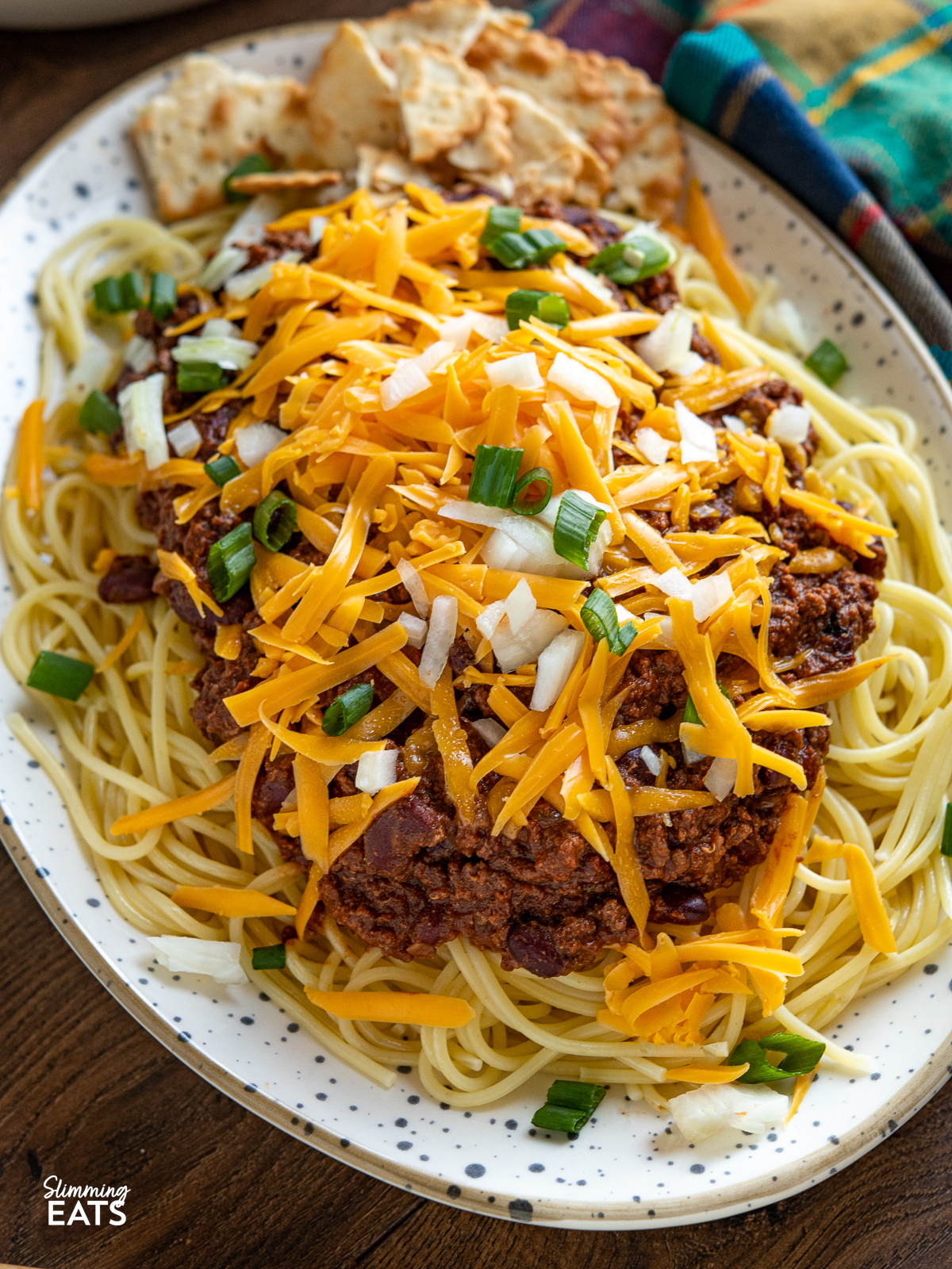 close up of Cincinnati Chili on spotted oval plate with spaghetti and toppings of cheese, onions and salted crackers