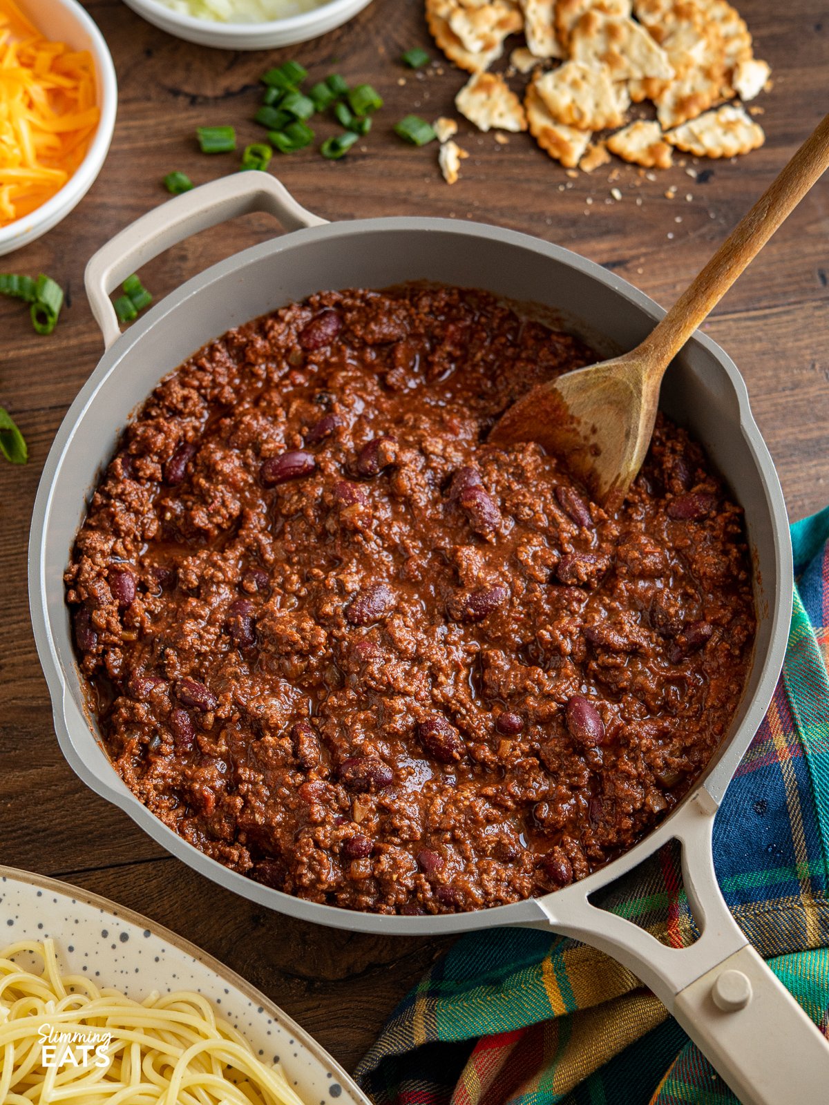 Cincinnati Chili in frying pan with wooden spoon, sides and toppings place around pan