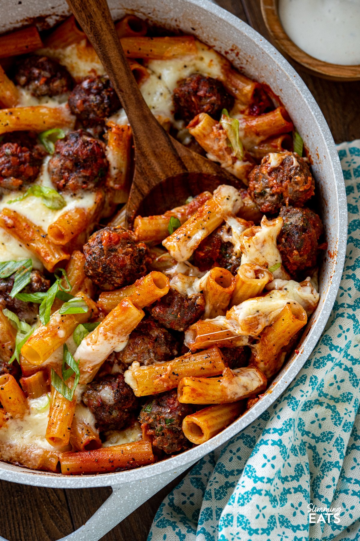 close up of pasta and meatballs being scooped from ceramic coated pan with wooden spoon showing the pull of melted mozzarella