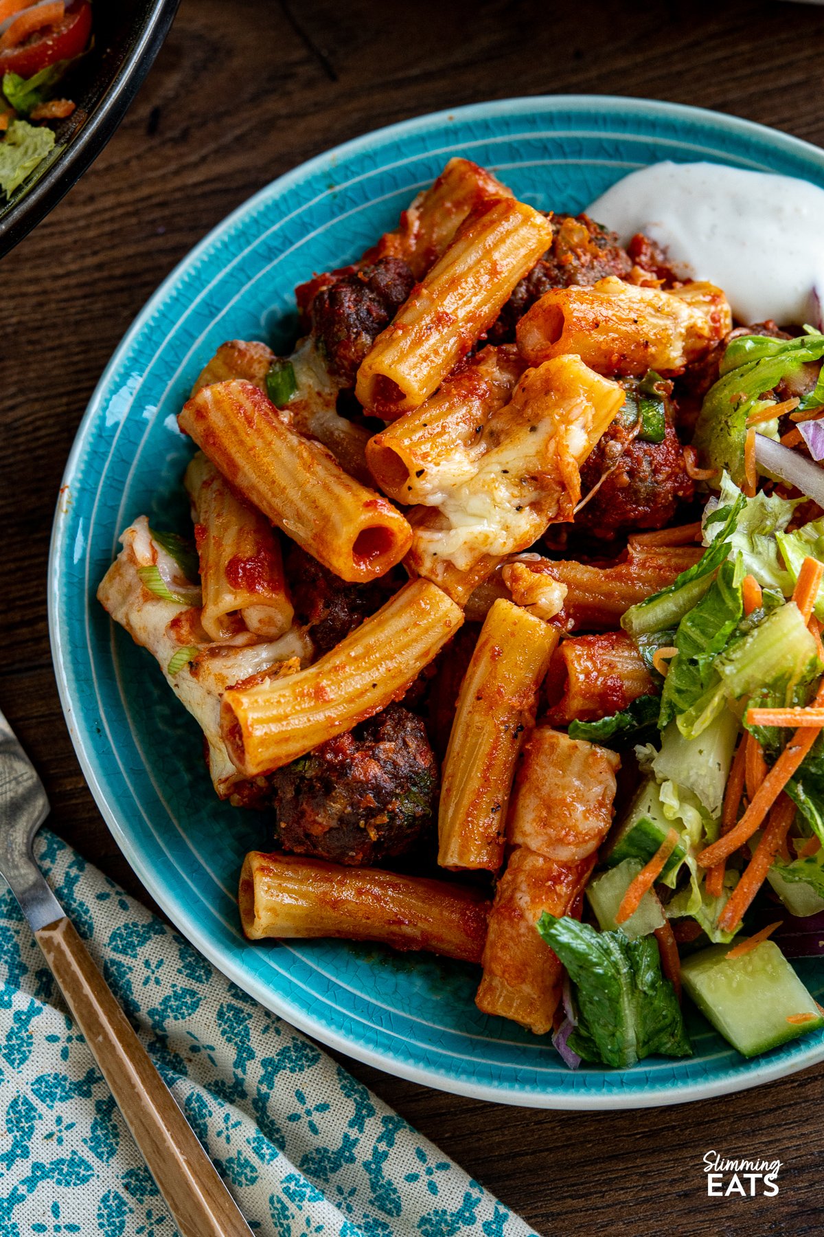Indulge in the ultimate comfort food with this delicious Honey Buffalo Meatball Pasta Bake. Featuring extra lean ground beef, sautéed vegetables in place of breadcrumbs, and a lower calorie homemade honey buffalo sauce, this recipe takes a classic dish to new heights. Get ready to satisfy your cravings with a flavour-packed and melted cheesy-topped delight that will leave you craving more close up of serving of Honey Buffalo Meatball Pasta Bake on turquoise plate with salad and ranch dressing