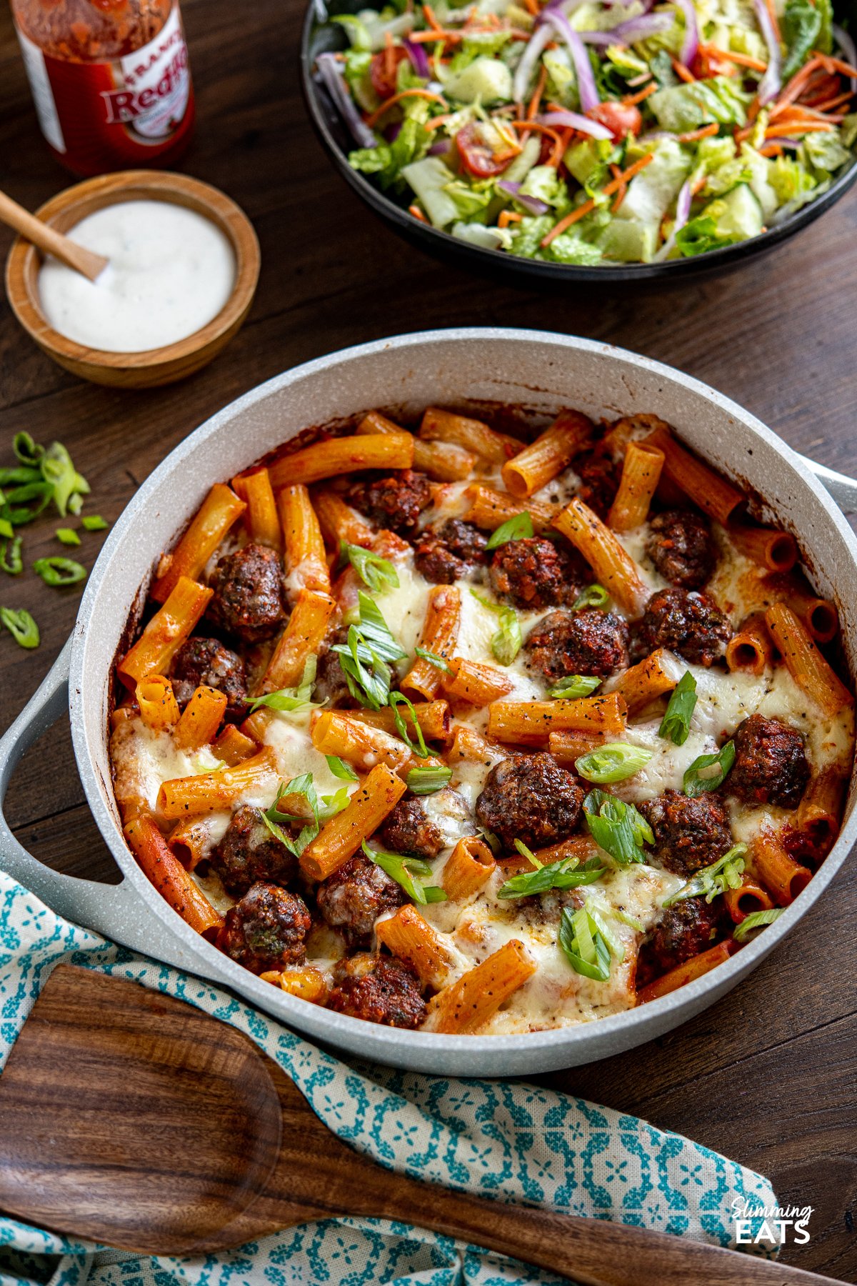 Indulge in the ultimate comfort food with this delicious Honey Buffalo Meatball Pasta Bake. Featuring extra lean ground beef, sautéed vegetables in place of breadcrumbs, and a lower calorie homemade honey buffalo sauce, this recipe takes a classic dish to new heights. Get ready to satisfy your cravings with a flavour-packed and melted cheesy-topped delight that will leave you craving more meatballs with pasta and melted mozzarella topping in ceramic coated pan with side salad in black bowl