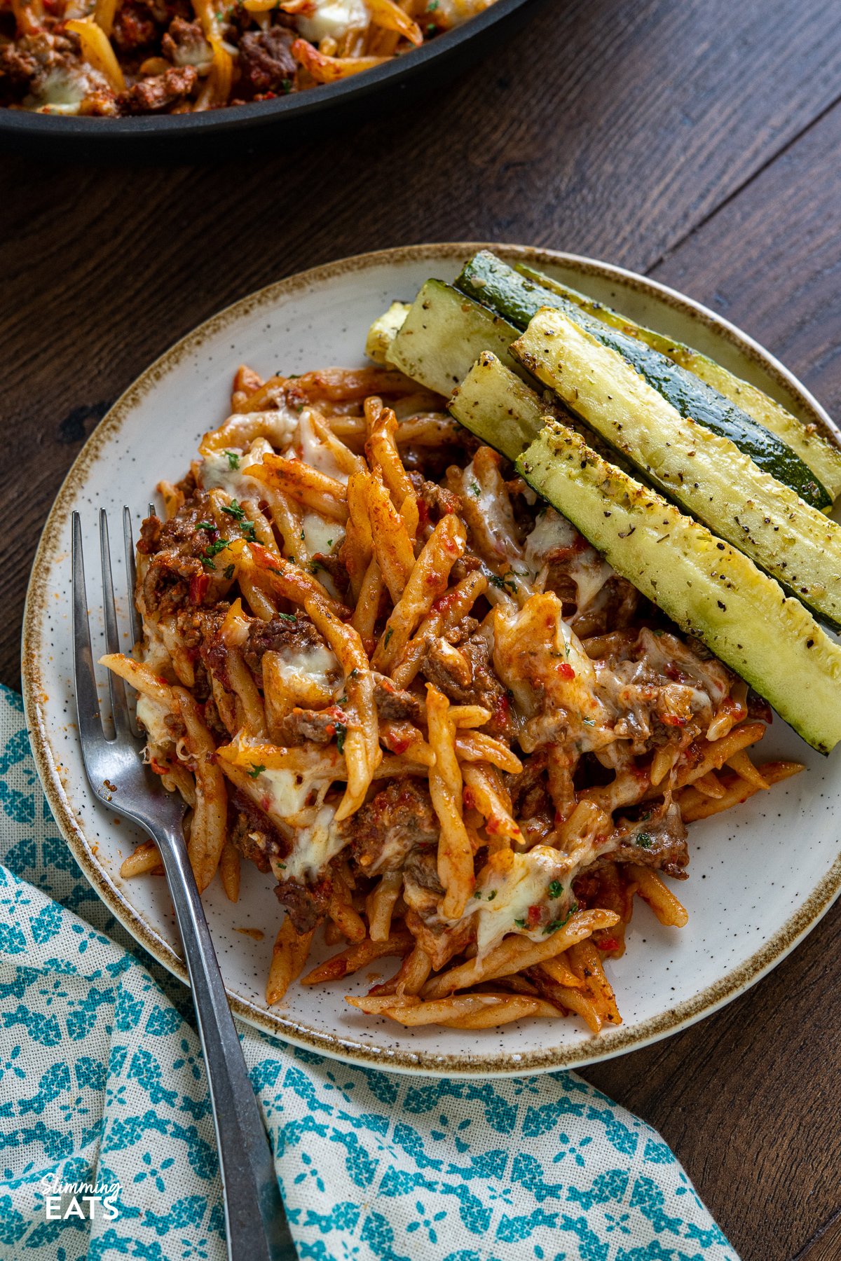 Creamy Beef Roasted Red Pepper Pasta Bake - Indulge in the ultimate comfort food with our creamy ground beef pasta recipe. This delectable dish features trofie pasta, roasted red peppers, and a heavenly blend of torn mozzarella and Parmesan. Baked to perfection, each bite is a symphony of flavors that will leave you craving more. Get ready to experience a mouthwatering culinary delight like no other. serving of Creamy Beef Roasted Red Pepper Pasta Bake with roasted zucchini on a white plate with beige rim fork placed to the left