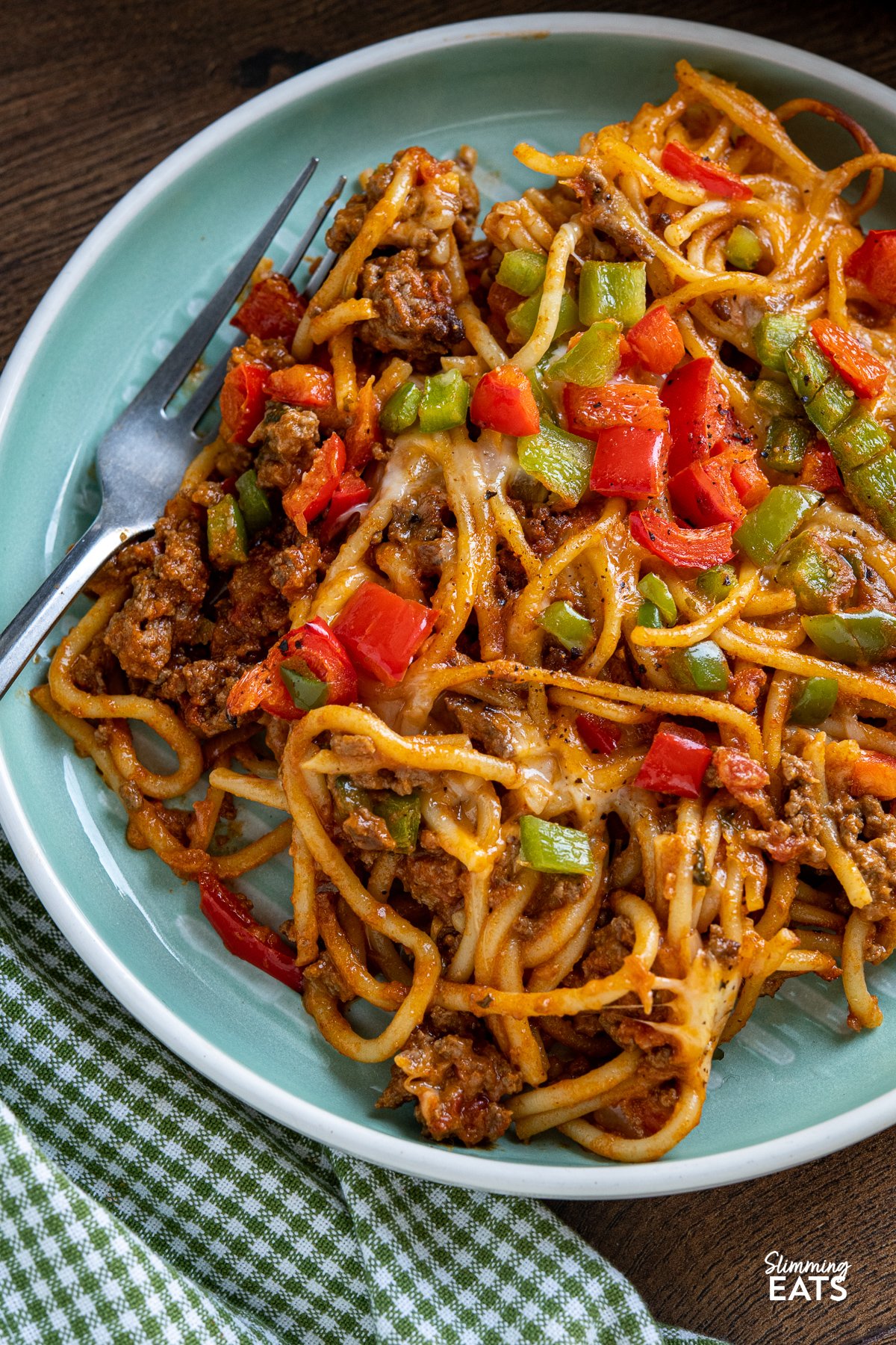 Baked Cheesy Taco SpaghettI - flavourful taco meat sauce, tangy salsa, and al dente spaghetti baked together to perfection, topped with a generous layer of gooey melted cheese and colorful bell peppers. It's a delightful blend of Tex-Mex flavors that will instantly become a family favourite. close up of serving of taco spaghetti bake in white oven proof dish on turquoise and white plate with fork to left