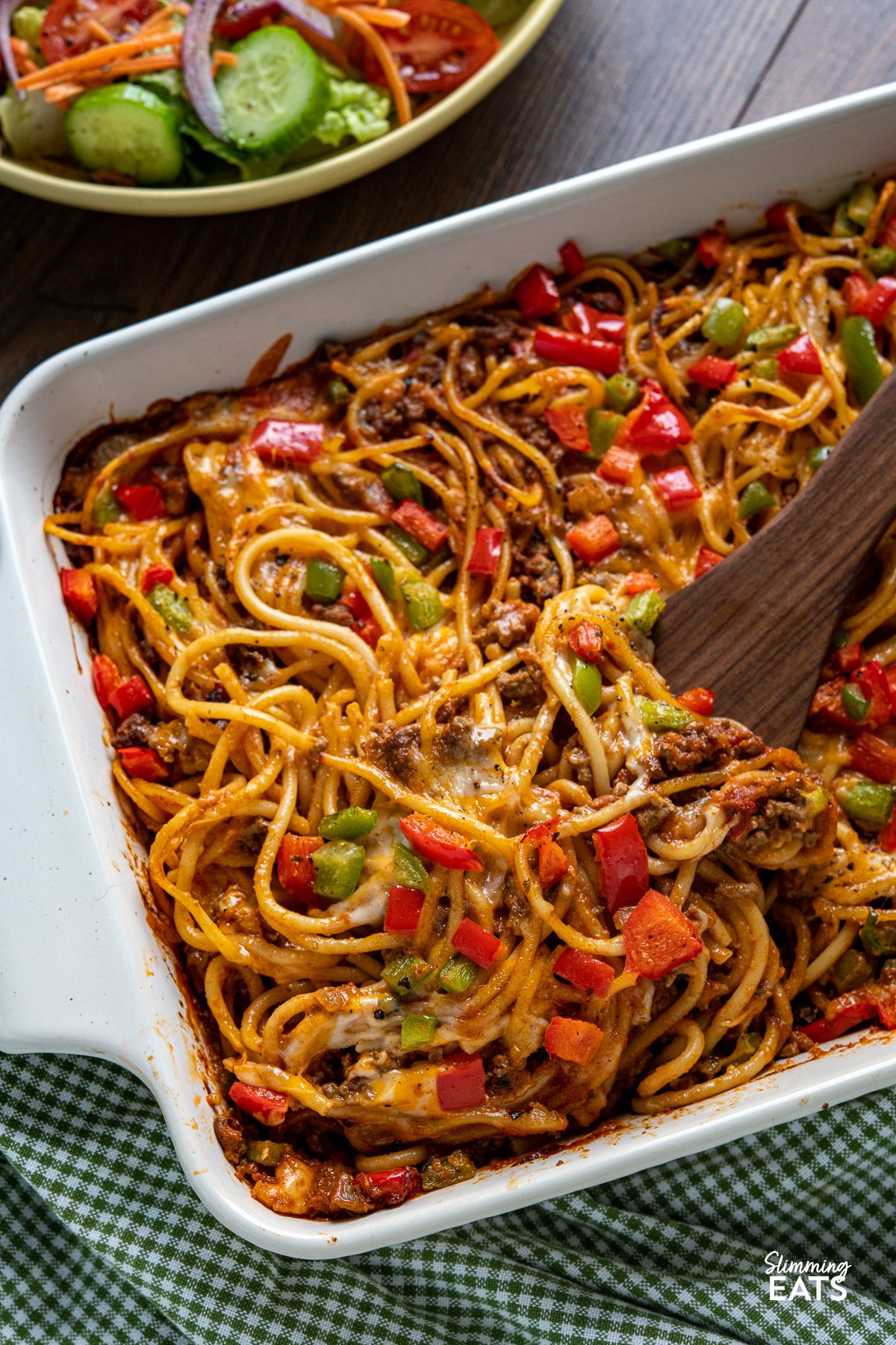 Baked Cheesy Taco SpaghettI - flavourful taco meat sauce, tangy salsa, and al dente spaghetti baked together to perfection, topped with a generous layer of gooey melted cheese and colorful bell peppers. It's a delightful blend of Tex-Mex flavors that will instantly become a family favourite. wooden spatula scooping portion of taco spaghetti bake from white oven proof dish