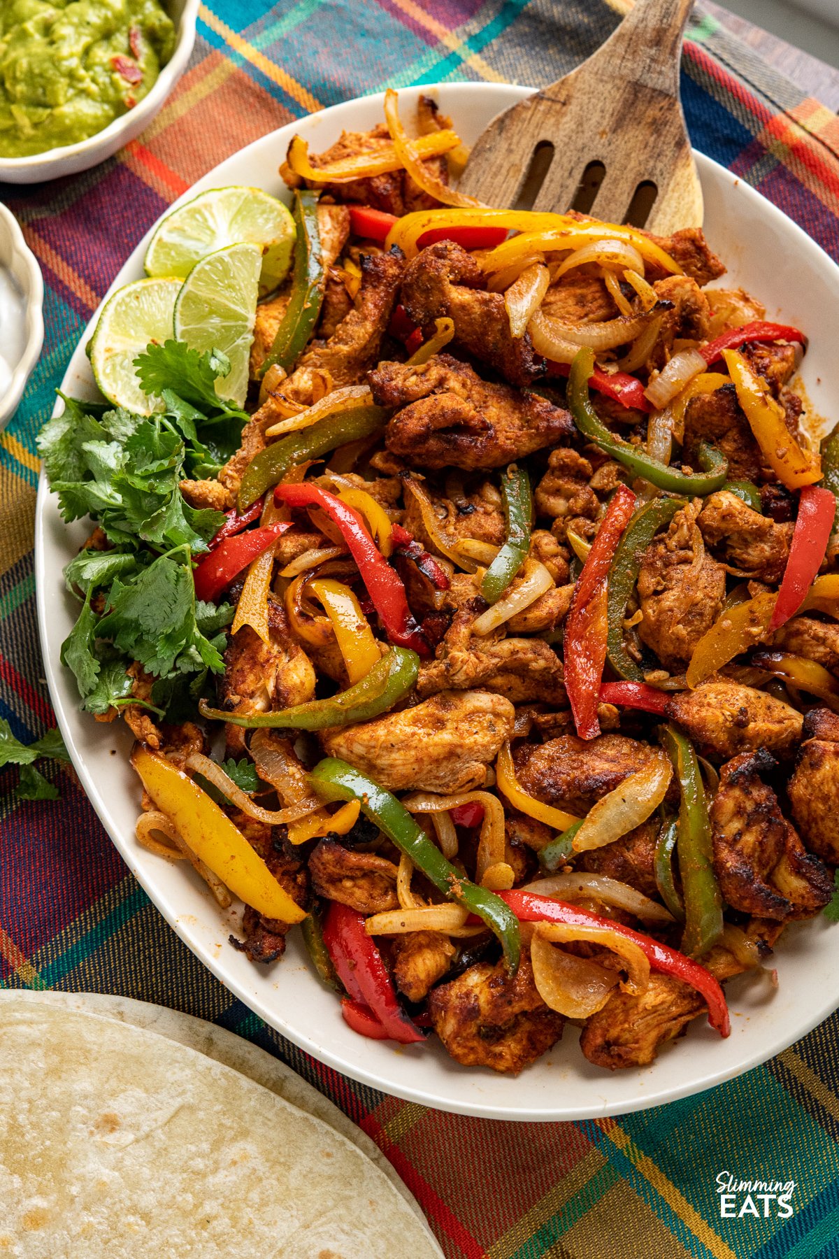 close up of Air Fryer Chicken Fajitas on a large oval plate with small bowls of guacamole and soured cream to the side.