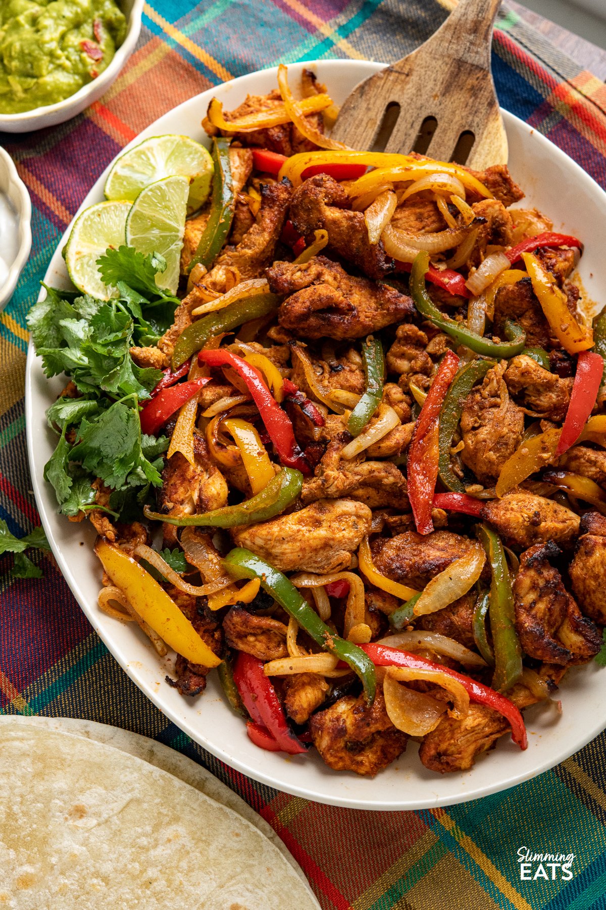 Are you looking for a quick and easy delicious dinner that is perfect for the whole family? Look no further than these simple Air Fryer Chicken Fajitas with tender chicken strips and mixed peppers and onion, all in the perfect homemade fajita seasoning blend. close up of Air Fryer Chicken Fajitas on a large oval plate with small bowls of guacamole and soured cream to the side.