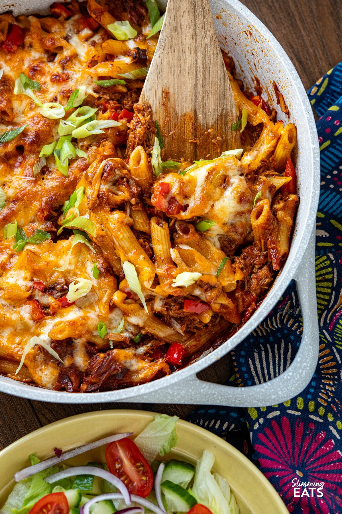 Pulled Pork Pasta Bake - combines the rich, smoky flavors of homemade leftover pulled pork with perfectly cooked pasta, all baked to cheesy, bubbling perfection. close up of Pulled Pork Pasta Bake in ceramic double handled light grey pan with yellow bowl of mixed salad