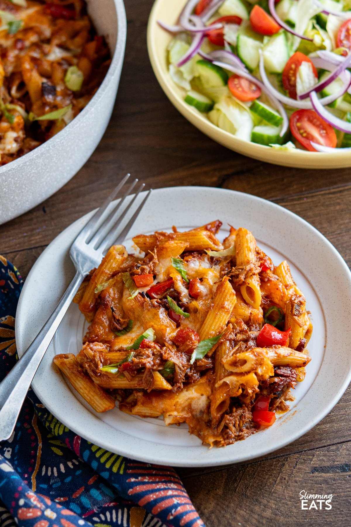 serving of Pulled Pork Pasta Bake on beige and white plate with silver fork with pan and bowl of salad in background