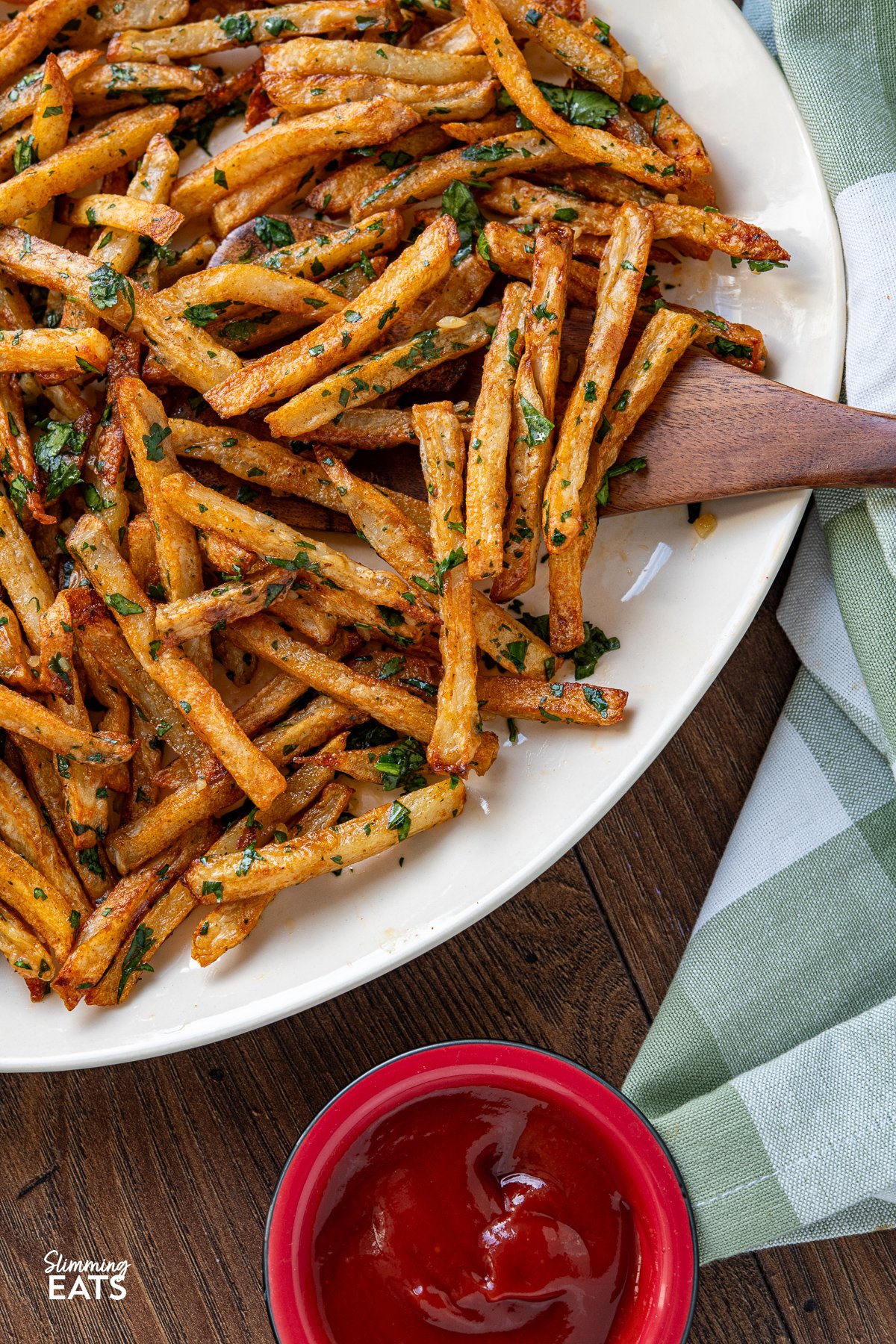 Savour the delicious fusion of these oven-baked seasoned fries, boasting a perfect crispiness, and the tantalizing harmony of garlic, butter, and cilantro. These delectable Garlic Cilantro Fries aren't just a side dish; they're a culinary revelation, ready to be relished either on their own with your preferred dip or as the star of any meal. close up of Garlic Cilantro Oven Baked Fries on oval cream plate with green and white checkered napkin