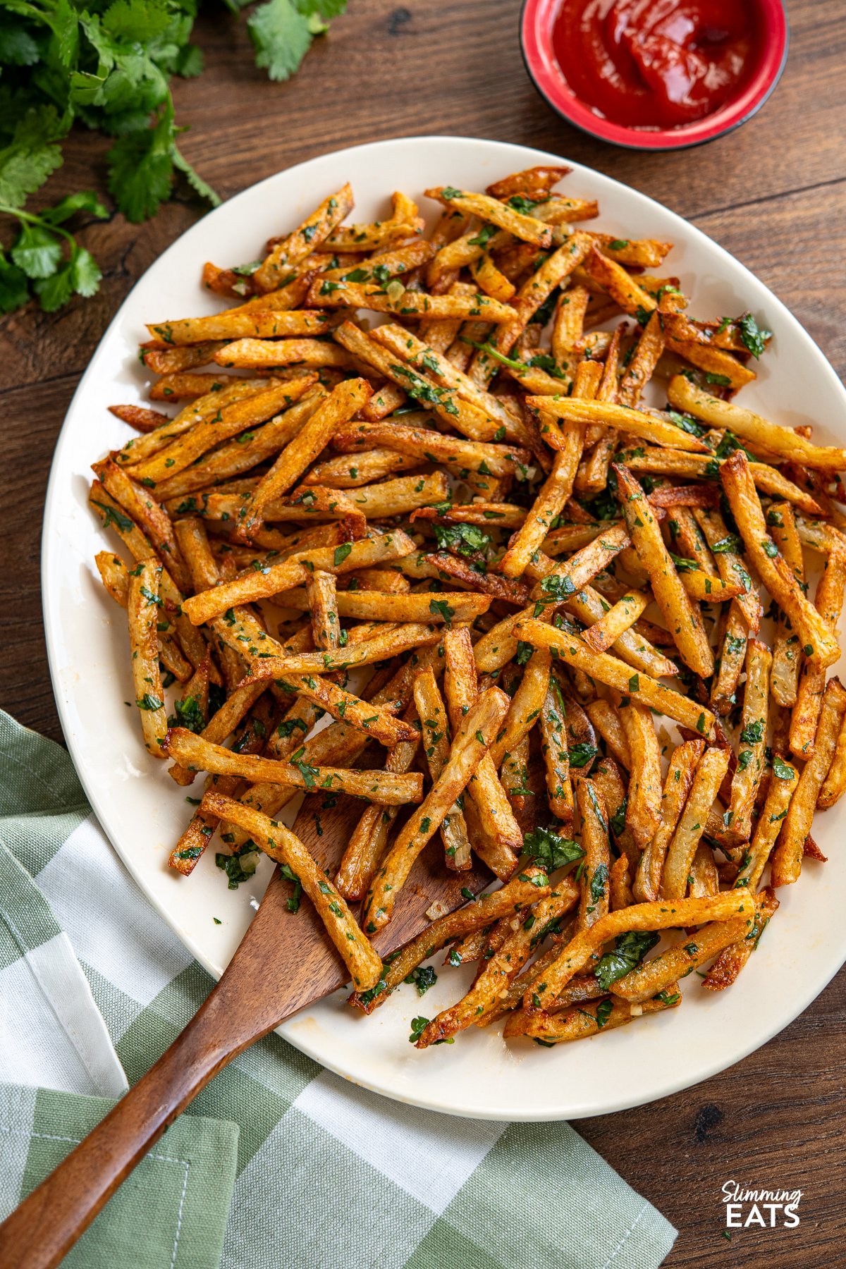 Savour the delicious fusion of these oven-baked seasoned fries, boasting a perfect crispiness, and the tantalizing harmony of garlic, butter, and cilantro. These delectable Garlic Cilantro Fries aren't just a side dish; they're a culinary revelation, ready to be relished either on their own with your preferred dip or as the star of any meal. Garlic Cilantro Oven Baked Fries on oval plate with wooden spatula and ketchup in small red bowl