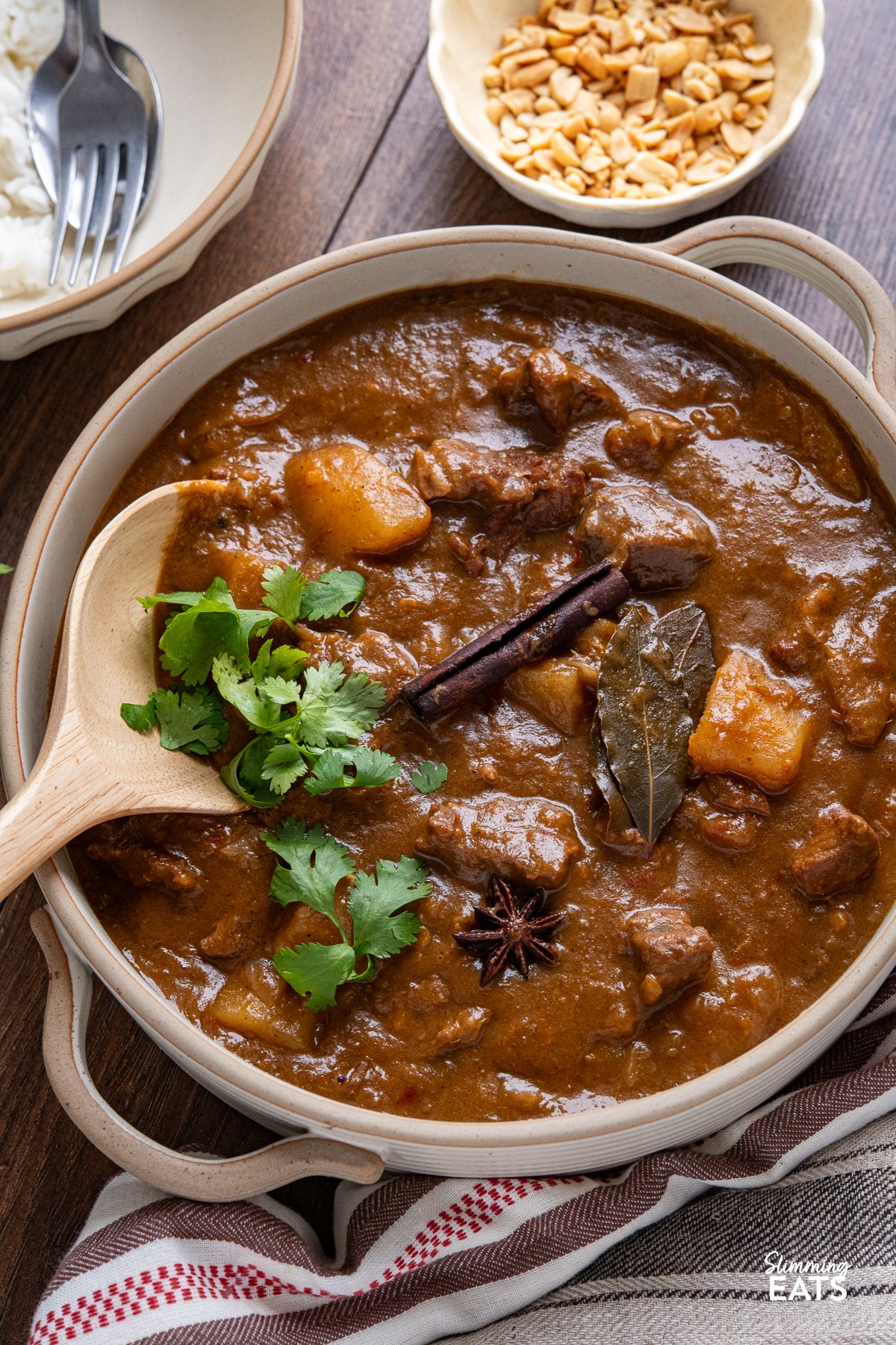 close up of Beef Massaman Curry in a beige ceramic pot with two handles bowl of rice above with peanuts in a small bowl.