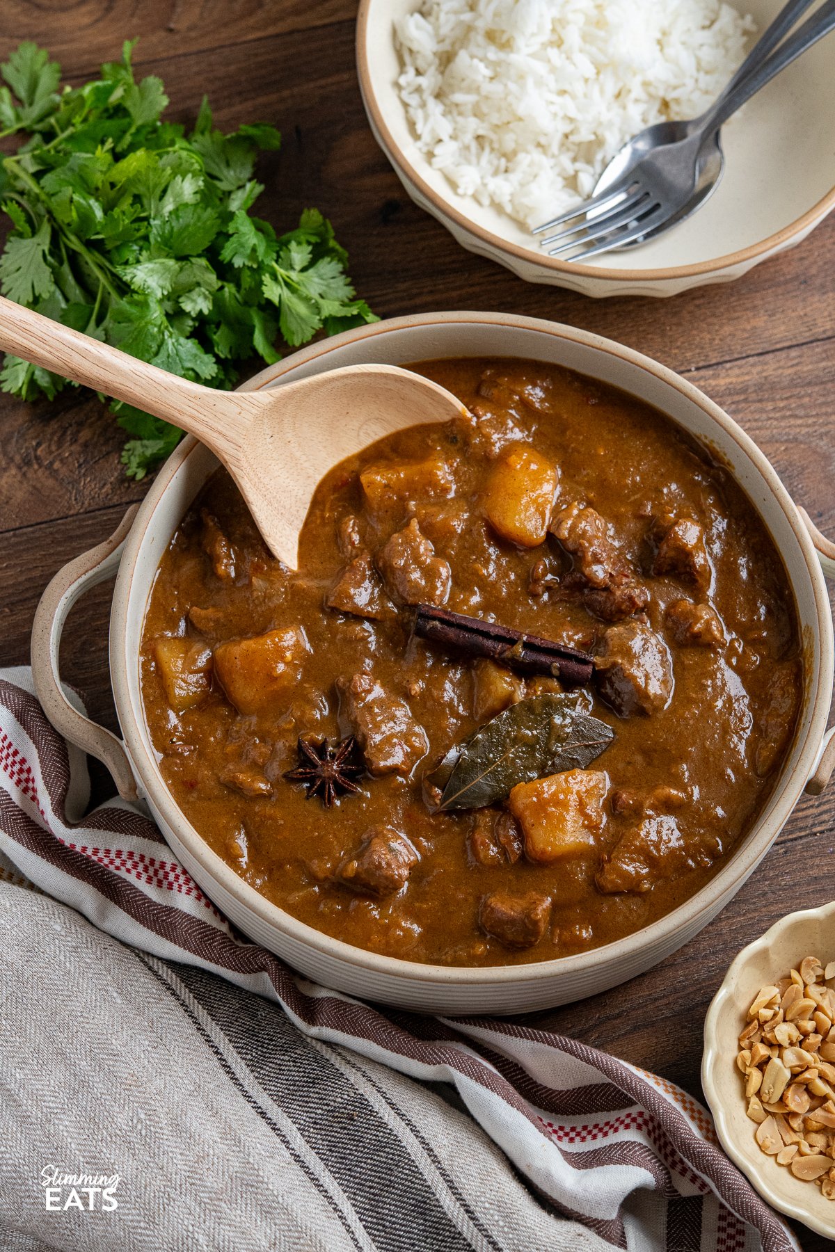 Beef Massaman Curry in a beige ceramic pot with two handles bowl of rice above with peanuts in a small bowl.