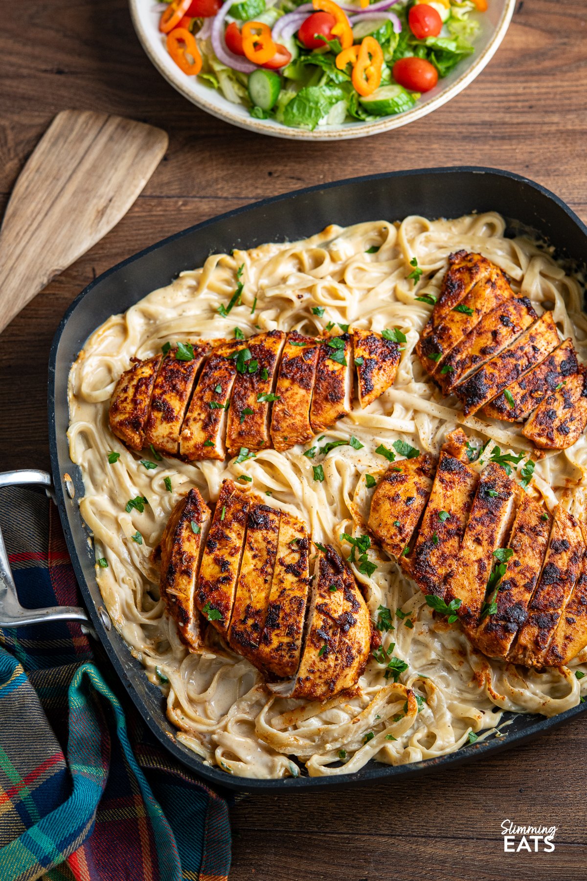 One Pot Cajun Chicken Alfredo - This mouthwatering dish features tender chicken breast seasoned to perfection with a homemade blend of bold Cajun spices, nestled into a lighter creamy Alfredo-coated fettuccine. One Pot Cajun Chicken Alfredo Pasta in a square frying pan with bowl of salad above.