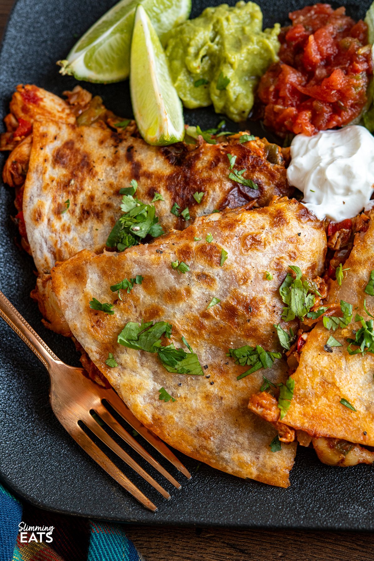 A serving of chicken quesadilla on a black plate, accompanied by lettuce, guacamole, salsa, and soured cream, decorated with scattered coriander and lime wedges.