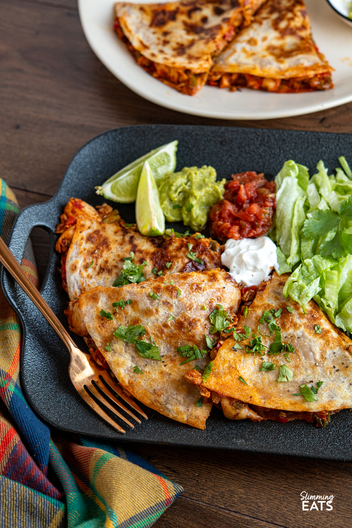 A serving of chicken quesadilla on a black plate, accompanied by lettuce, guacamole, salsa, and soured cream, decorated with scattered coriander and lime wedges.