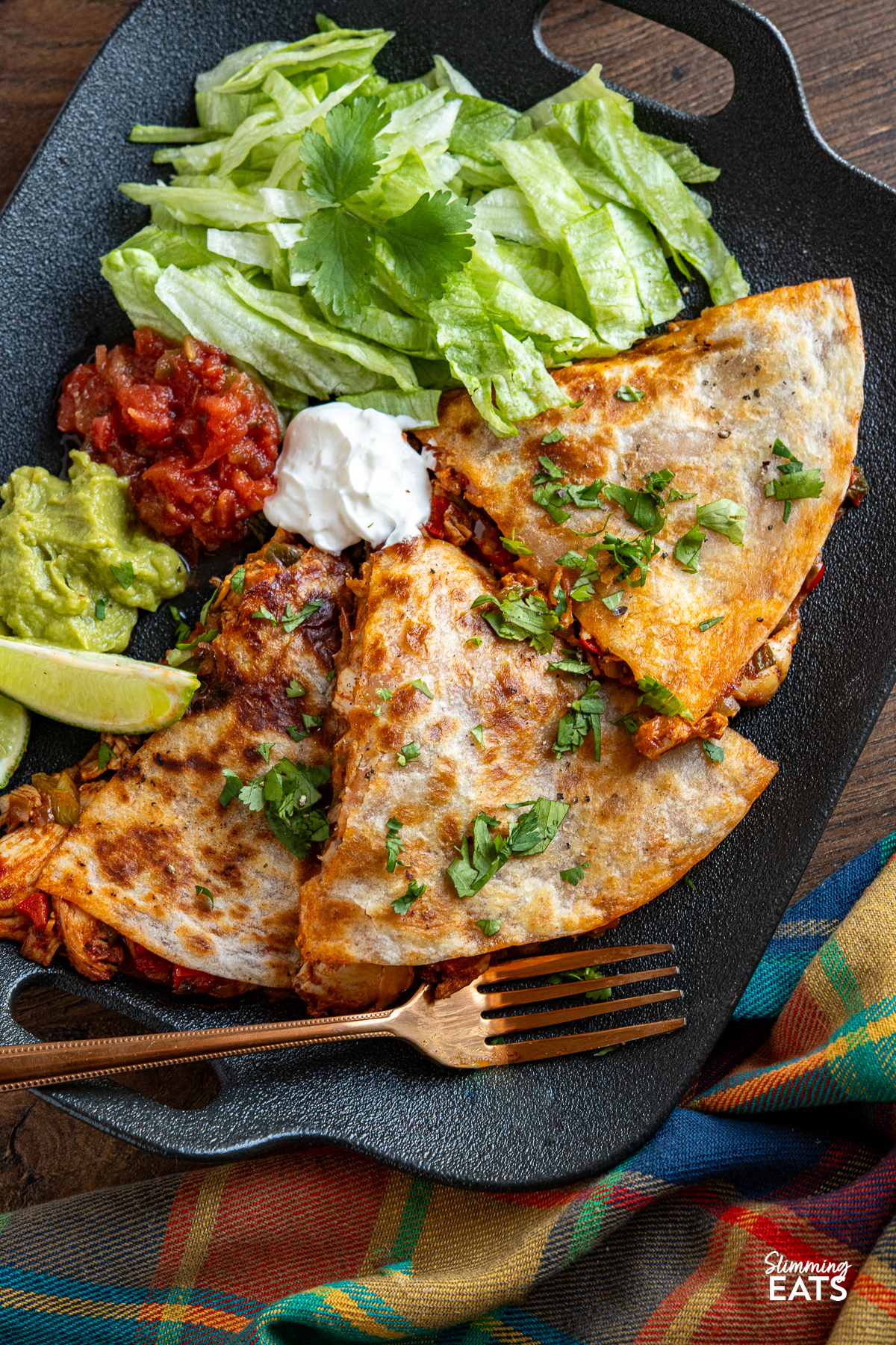 A serving of chicken quesadilla on a black plate, accompanied by lettuce, guacamole, salsa, and soured cream, decorated with scattered coriander and lime wedges.
