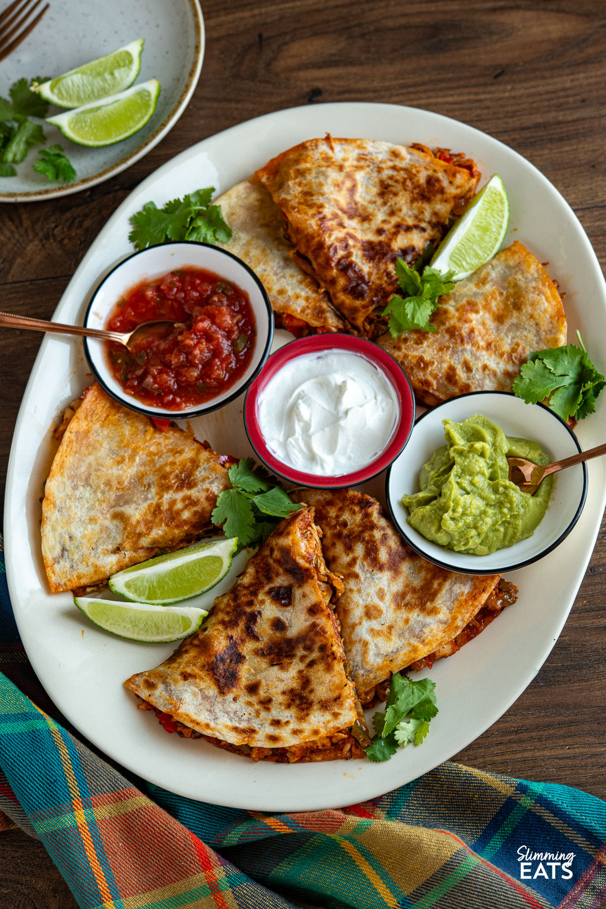 Sliced quesadilla arranged on a large cream oval plate, accompanied by small bowls of guacamole, salsa, and soured cream, garnished with scattered coriander leaves and lime wedges.