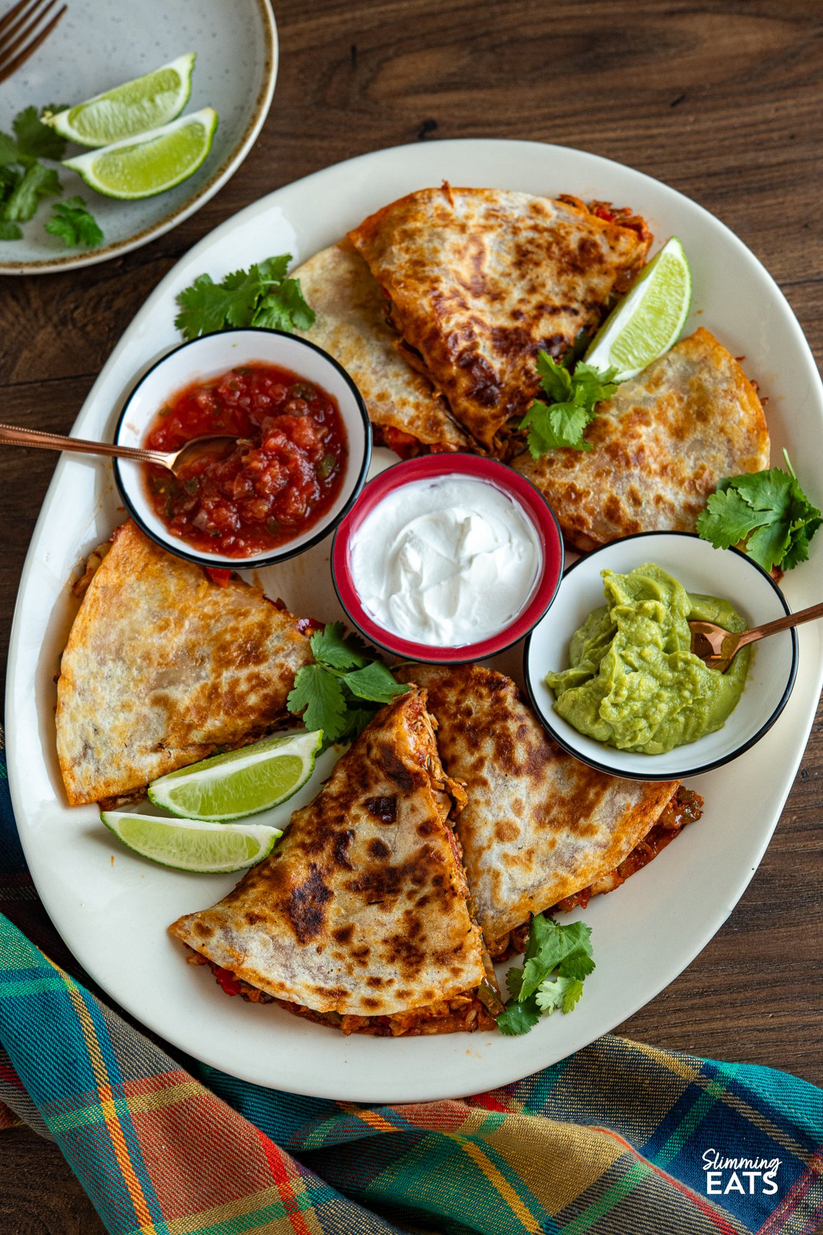 Sliced quesadilla arranged on a large cream oval plate, accompanied by small bowls of guacamole, salsa, and soured cream, garnished with scattered coriander leaves and lime wedges.