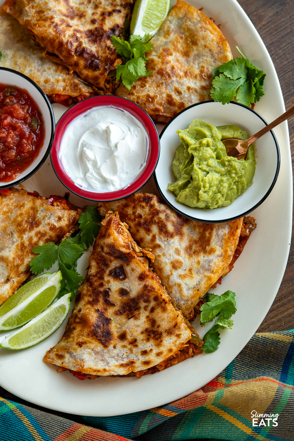 Sliced quesadilla arranged on a large cream oval plate, accompanied by small bowls of guacamole, salsa, and soured cream, garnished with scattered coriander leaves and lime wedges.