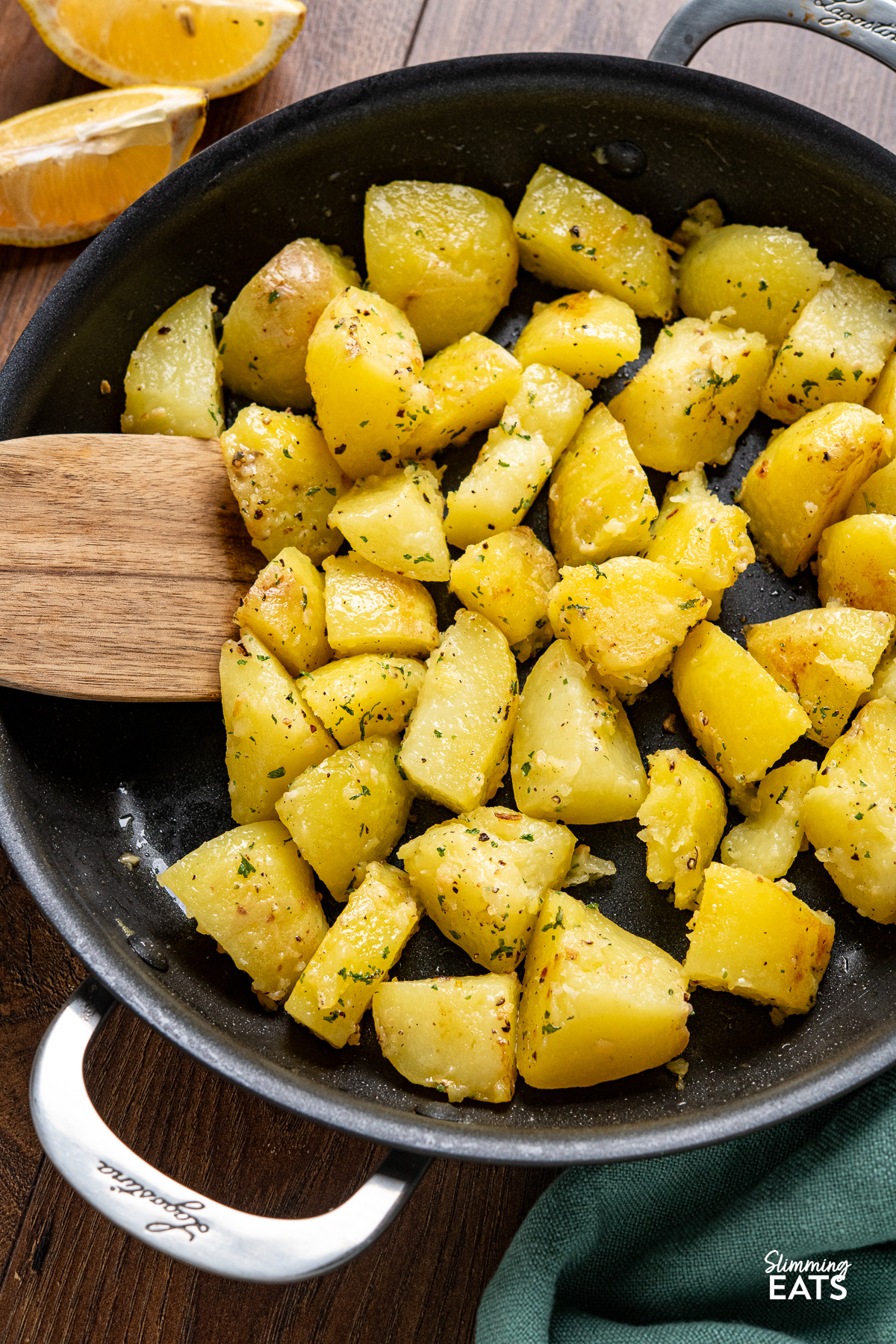 On a wooden board: Lemon garlic potatoes served in a non-stick double-handled skillet with wooden spatula and lemon slices