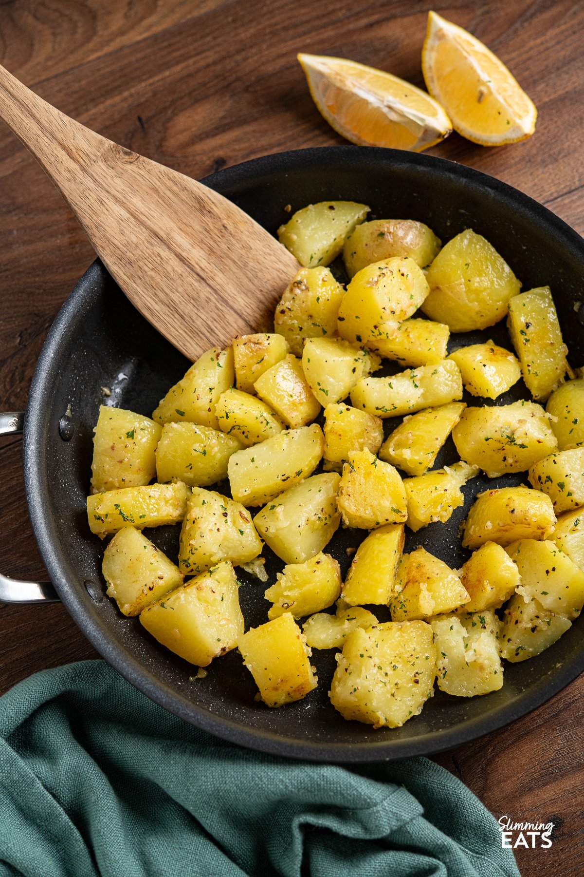 On a wooden board: Lemon garlic potatoes served in a non-stick double-handled skillet with wooden spatula and lemon slices