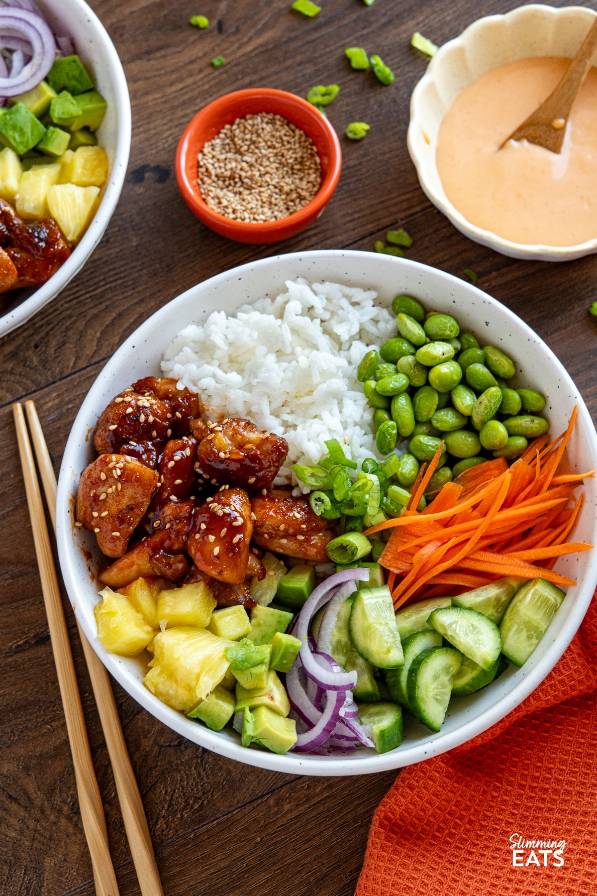White bowl filled with Hawaiian chicken poke bowl topped with assorted fresh toppings, accompanied by a small bowl of spicy mayo and small bowl of sesame seeds.