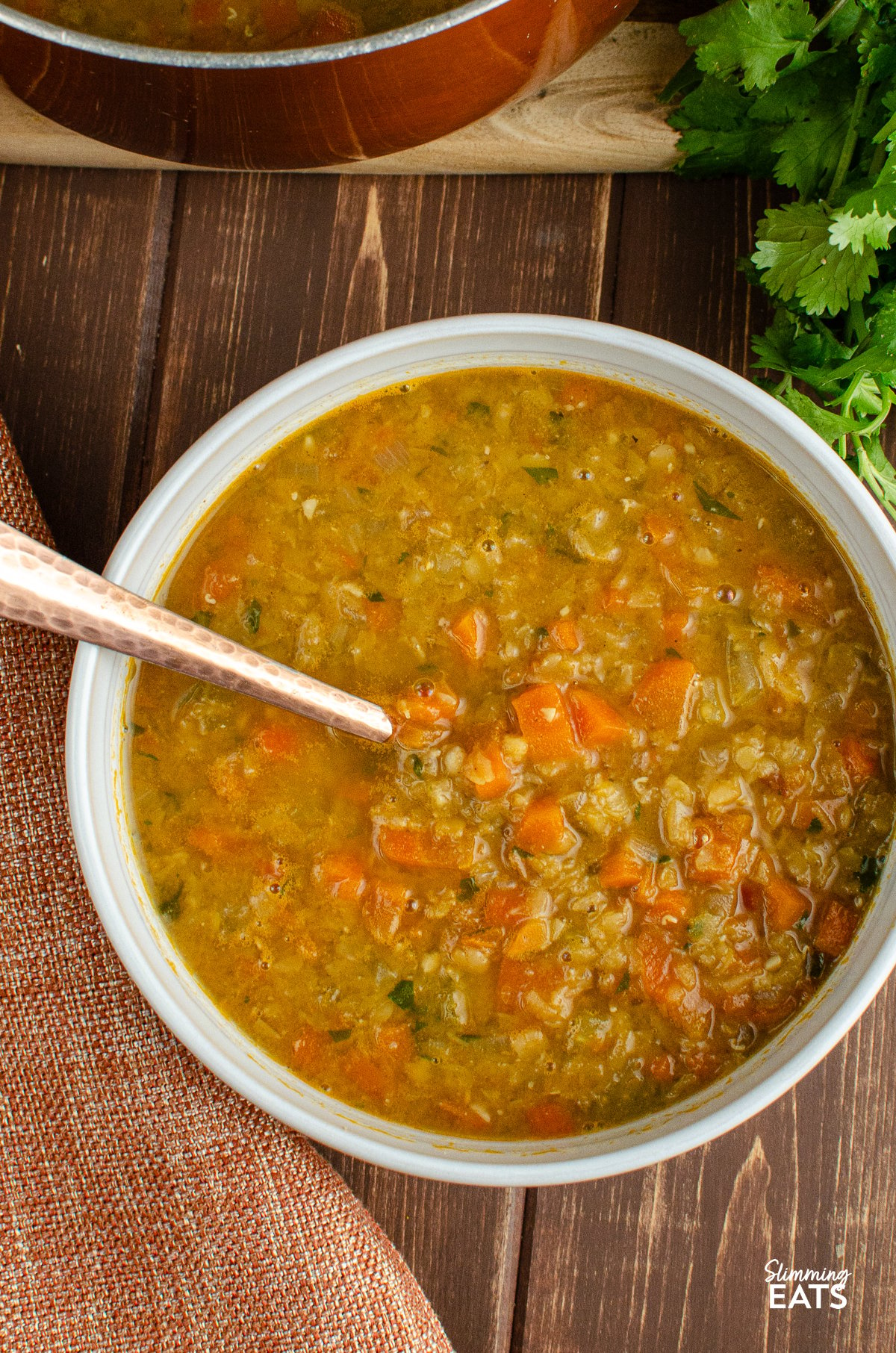 Spicy carrot and lentil soup served in a white bowl with a spoon, with the saucepan used for cooking visible in the background.