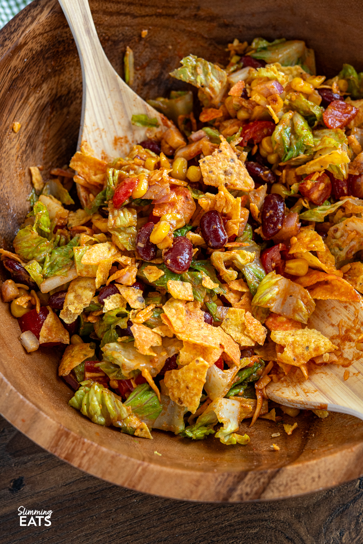 In a rustic wooden bowl, the Dorito Taco Salad is fully mixed, showcasing its vibrant colors and enticing aroma, inviting you to indulge with the aid of salad spoons.