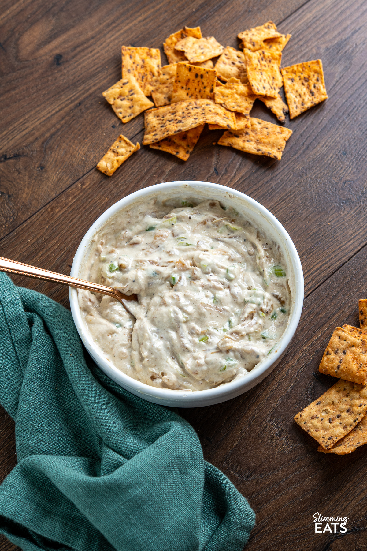 White bowl filled with creamy caramelized onion dip, garnished with a spoon, surrounded by scattered multigrain chips on wooden board