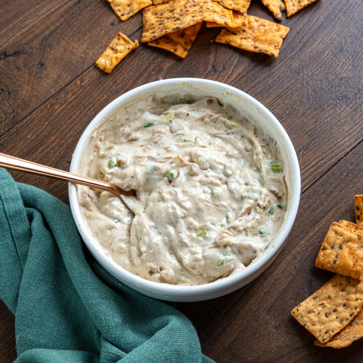 White bowl filled with creamy caramelized onion dip, garnished with a spoon, surrounded by scattered multigrain chips on wooden board