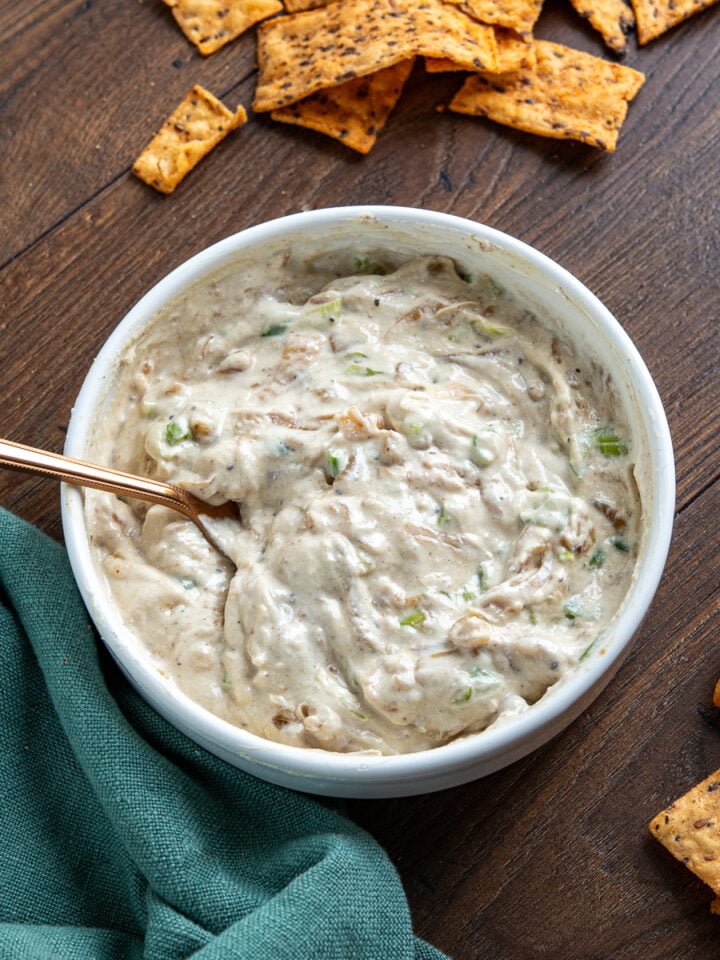 White bowl filled with creamy caramelized onion dip, garnished with a spoon, surrounded by scattered multigrain chips on wooden board