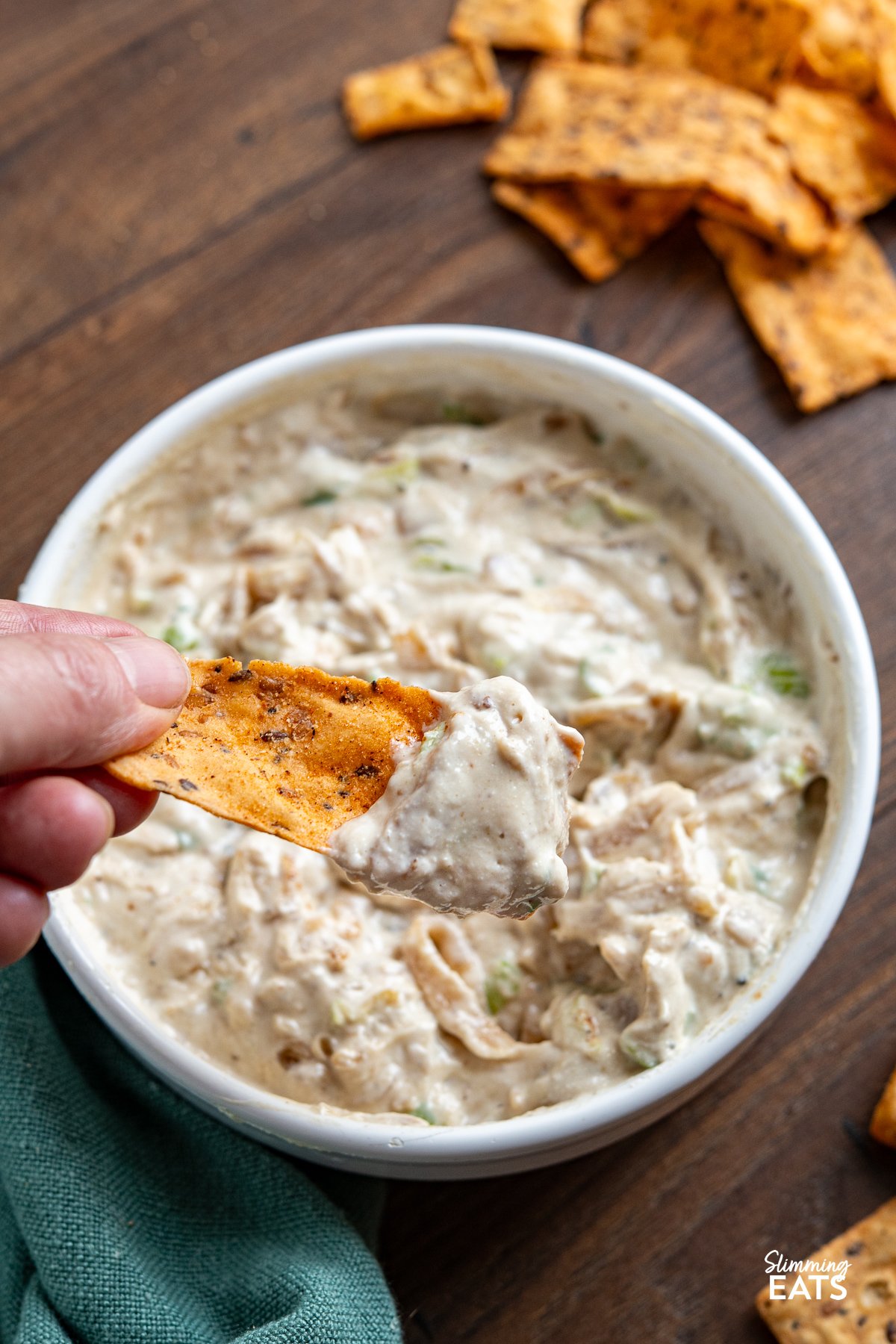 White bowl filled with creamy caramelized onion dip, garnished with a spoon, surrounded by scattered multigrain chips on wooden board