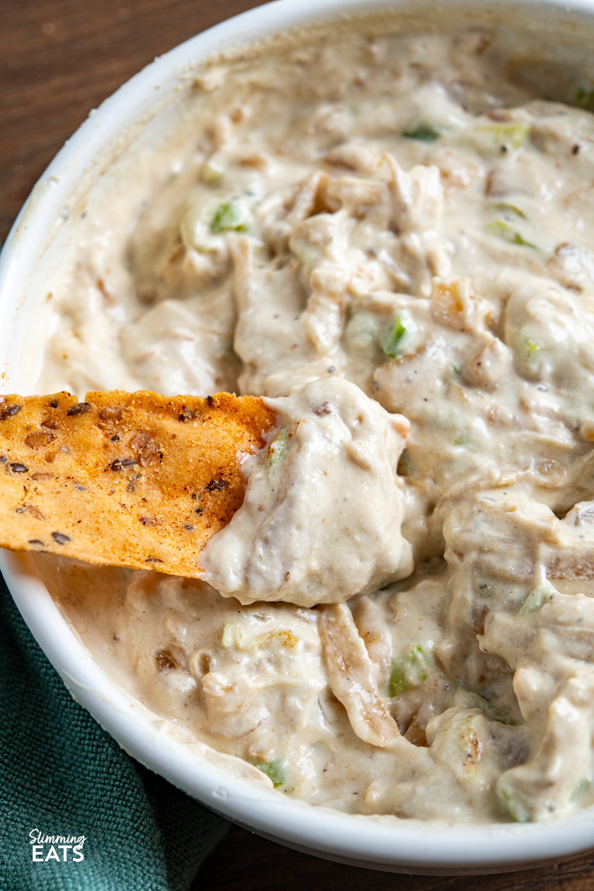 White bowl filled with creamy caramelized onion dip, garnished with a spoon, surrounded by scattered multigrain chips on wooden board