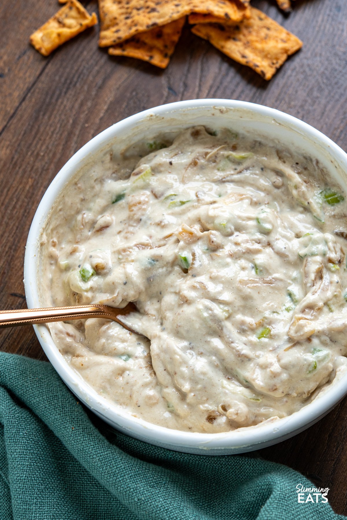 White bowl filled with creamy caramelized onion dip, garnished with a spoon, surrounded by scattered multigrain chips on wooden board