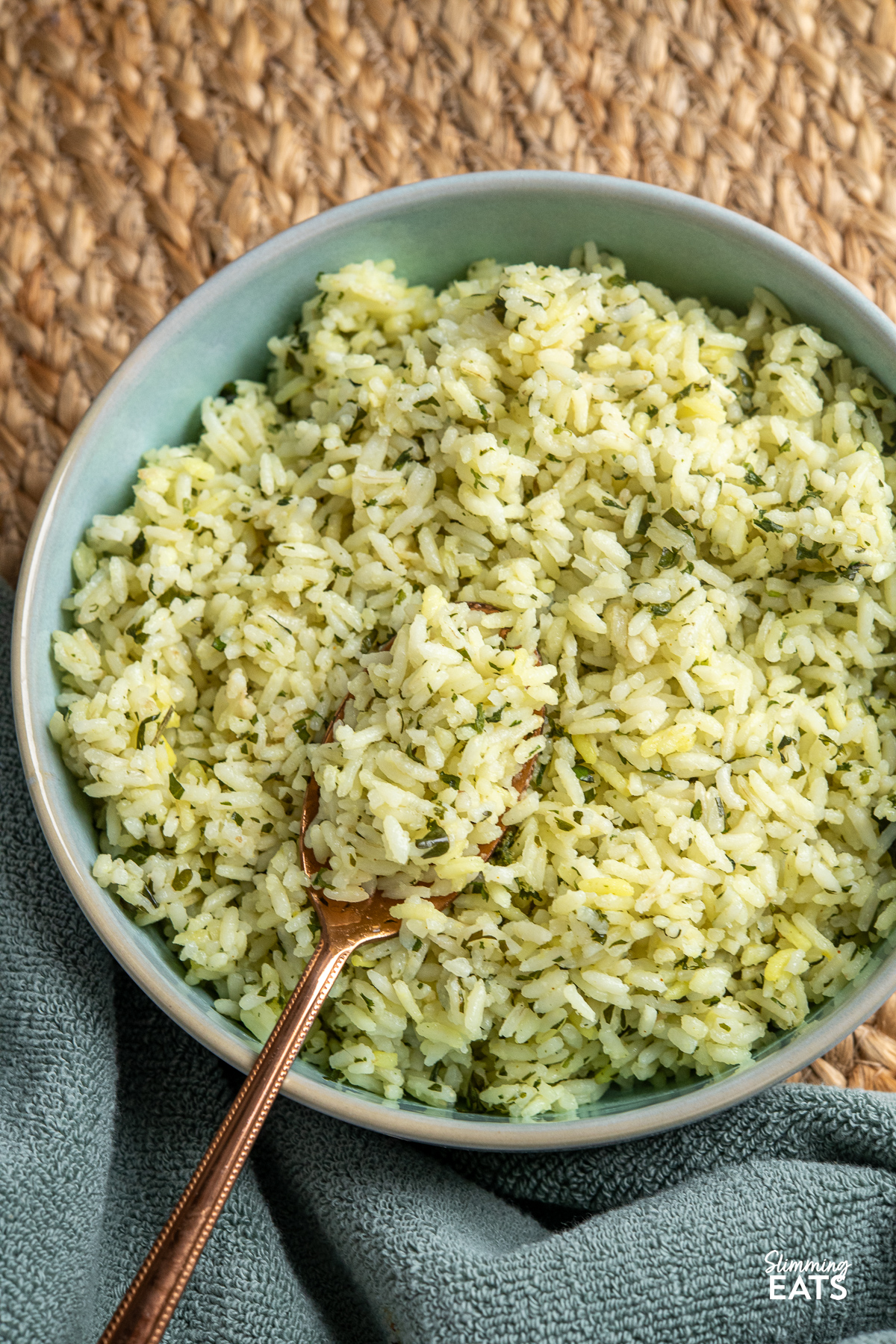 close up of Cooked herby garlic rice in light turquoise bowl with spoon placed on a mat.