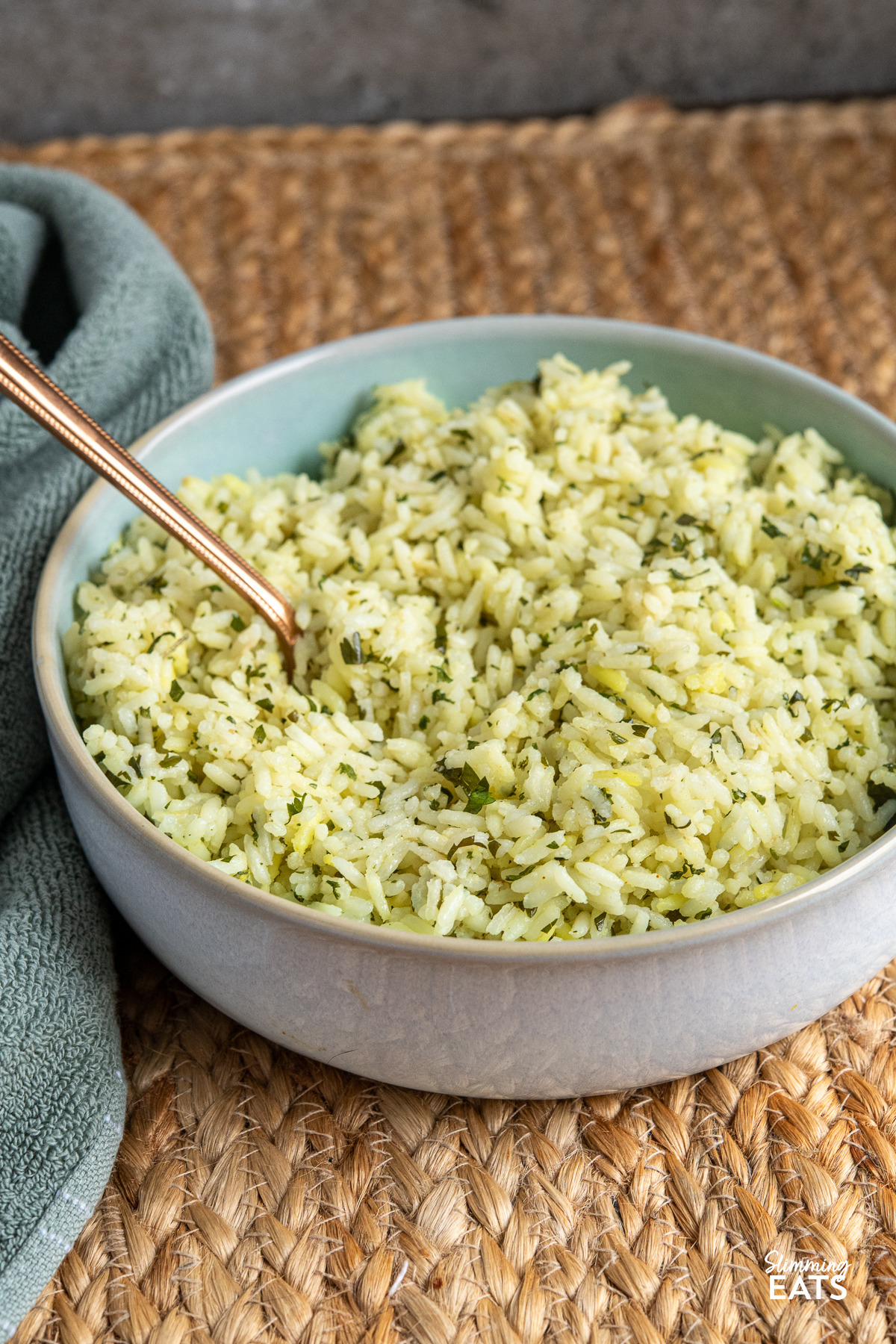 Cooked herby garlic rice in light turquoise bowl with spoon placed on a mat.