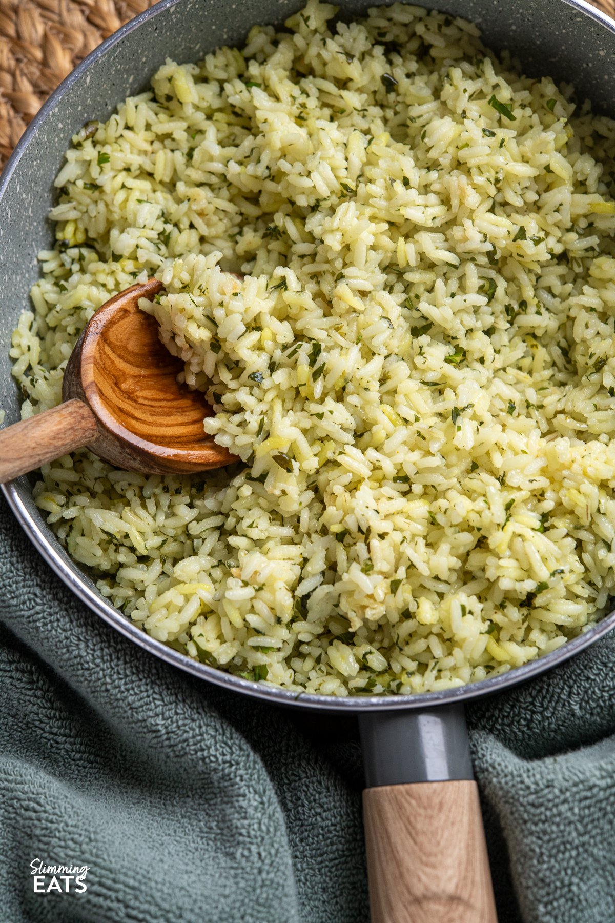 close up of Cooked herby garlic rice in a wooden handled saucepan with olive wood spoon placed in rice.