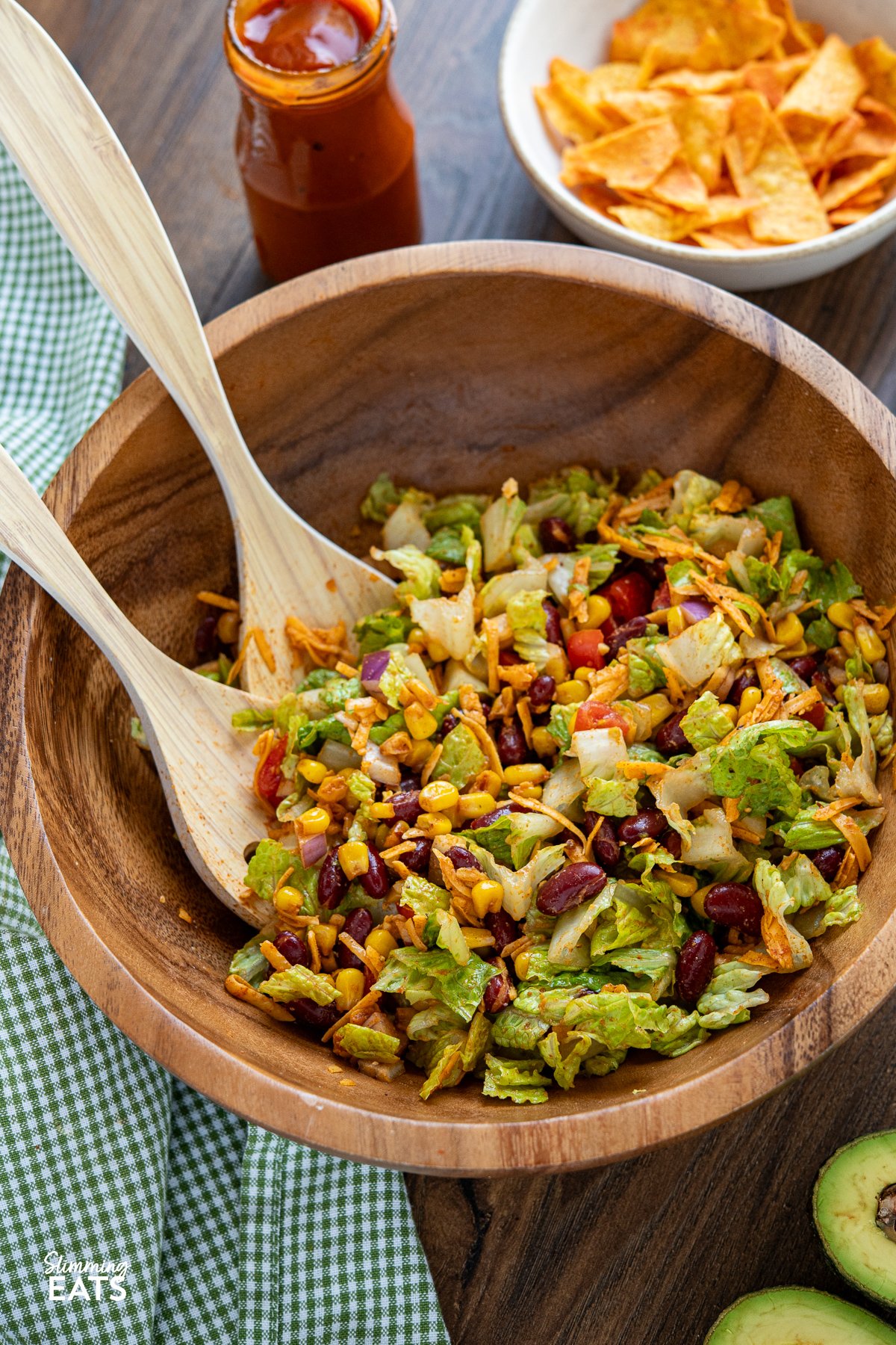 A wooden bowl holds a colourful medley of ingredients for Dorito Taco Salad, including lettuce, tomatoes, onions, and cheese, ready to be tossed together with salad spoons for a satisfying meal.