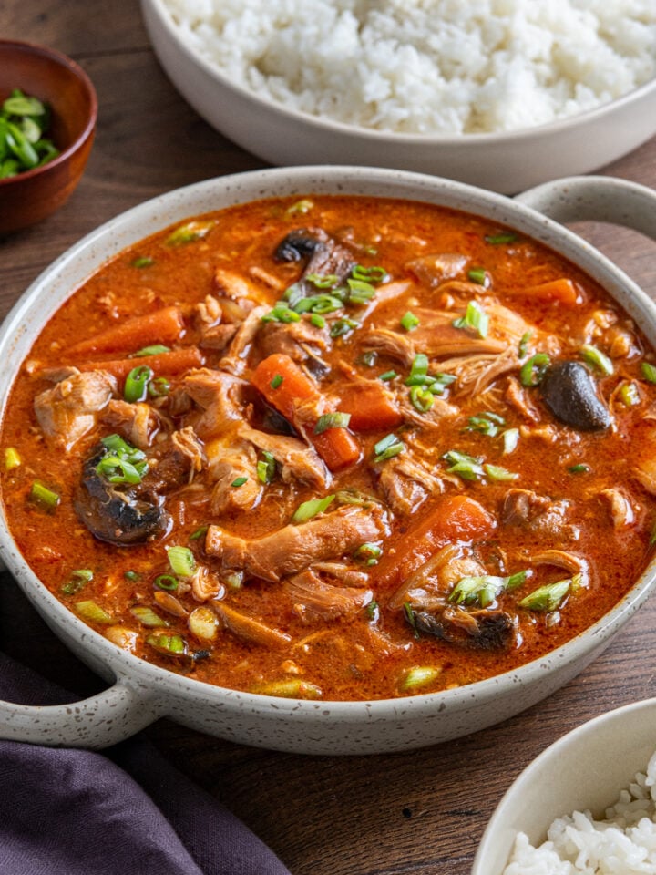 A double-handled ceramic dish filled with Korean Chicken Stew, with a bowl of sticky rice in the background and a small bowl of chopped green onions.