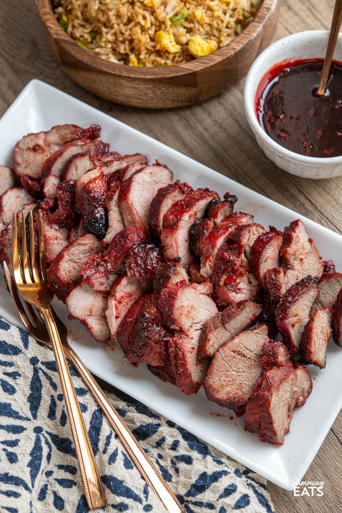 Easy Make-at-Home Char Siu Pork - Tender slices of pork, baked to perfection in the oven, are infused with the rich, tantalising flavours of the Chinese-style BBQ marinade. Sliced Char Siu Pork arranged on a white rectangular plate with two forks placed on the side. A bowl of egg fried rice and extra sauce are positioned above.
