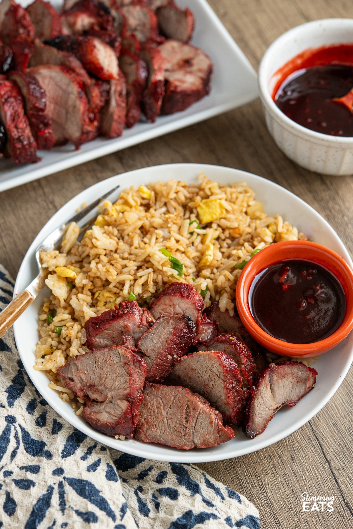 Easy Make-at-Home Char Siu Pork - Tender slices of pork, baked to perfection in the oven, are infused with the rich, tantalising flavours of the Chinese-style BBQ marinade. Sliced Char Siu Pork in a white bowl with egg fried rice and extra sauce, extra sauce and pork in background.