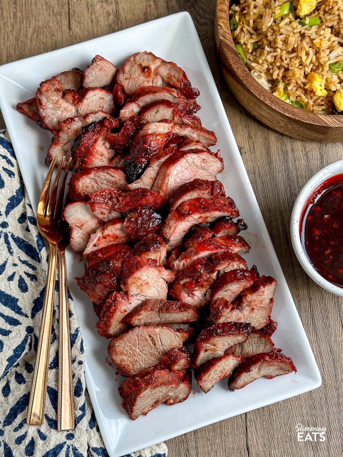Easy Make-at-Home Char Siu Pork - Tender slices of pork, baked to perfection in the oven, are infused with the rich, tantalising flavours of the Chinese-style BBQ marinade. Sliced Char Siu Pork arranged on a white rectangular plate with two forks placed on the side. A bowl of egg fried rice and extra sauce are positioned to the right.