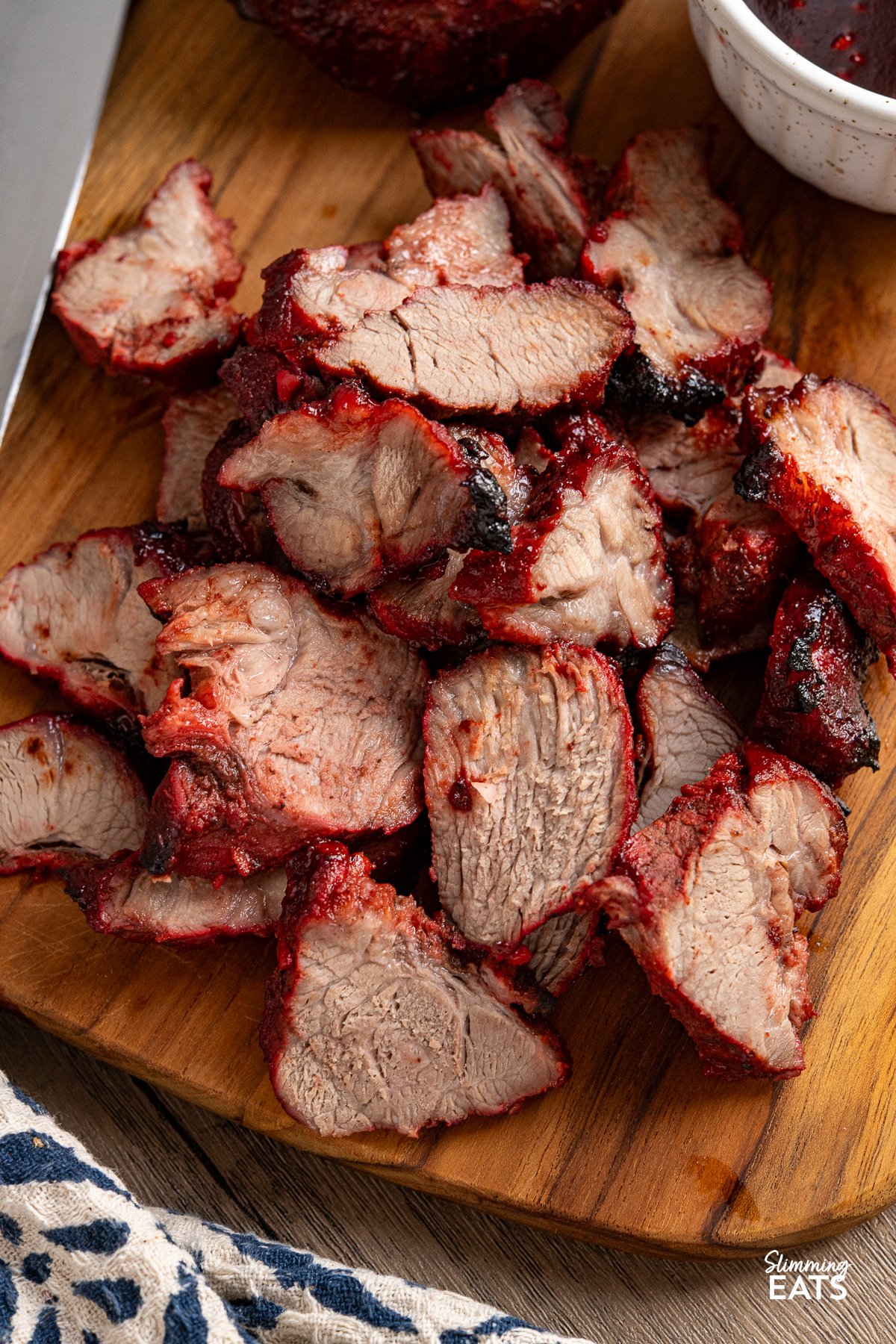 Easy Make-at-Home Char Siu Pork - Tender slices of pork, baked to perfection in the oven, are infused with the rich, tantalising flavours of the Chinese-style BBQ marinade. close up of char sui pork with knife of wooden board sliced with bowl of extra sauce on side