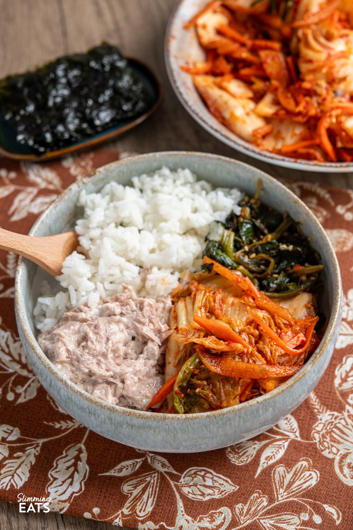 Looking to add a burst of vibrant flavour to your meals without the strong tang of traditional fermented kimchi? This quick and easy fresh kimchi recipe is the perfect introduction. By skipping the fermentation process, you get a crunchy, refreshing side dish that pairs wonderfully with all your favourite Korean dishes. Made with crisp napa cabbage, crunchy carrots, green onions, and sweet Asian pear, this delightful kimchi offers a spicy yet lightly sweet taste that will soon become a staple at your table. quick fresh kimchi in a light blue bowl with rice, tuna mayo, seasoned spinach and nori sheets and bowl of kimchi in background