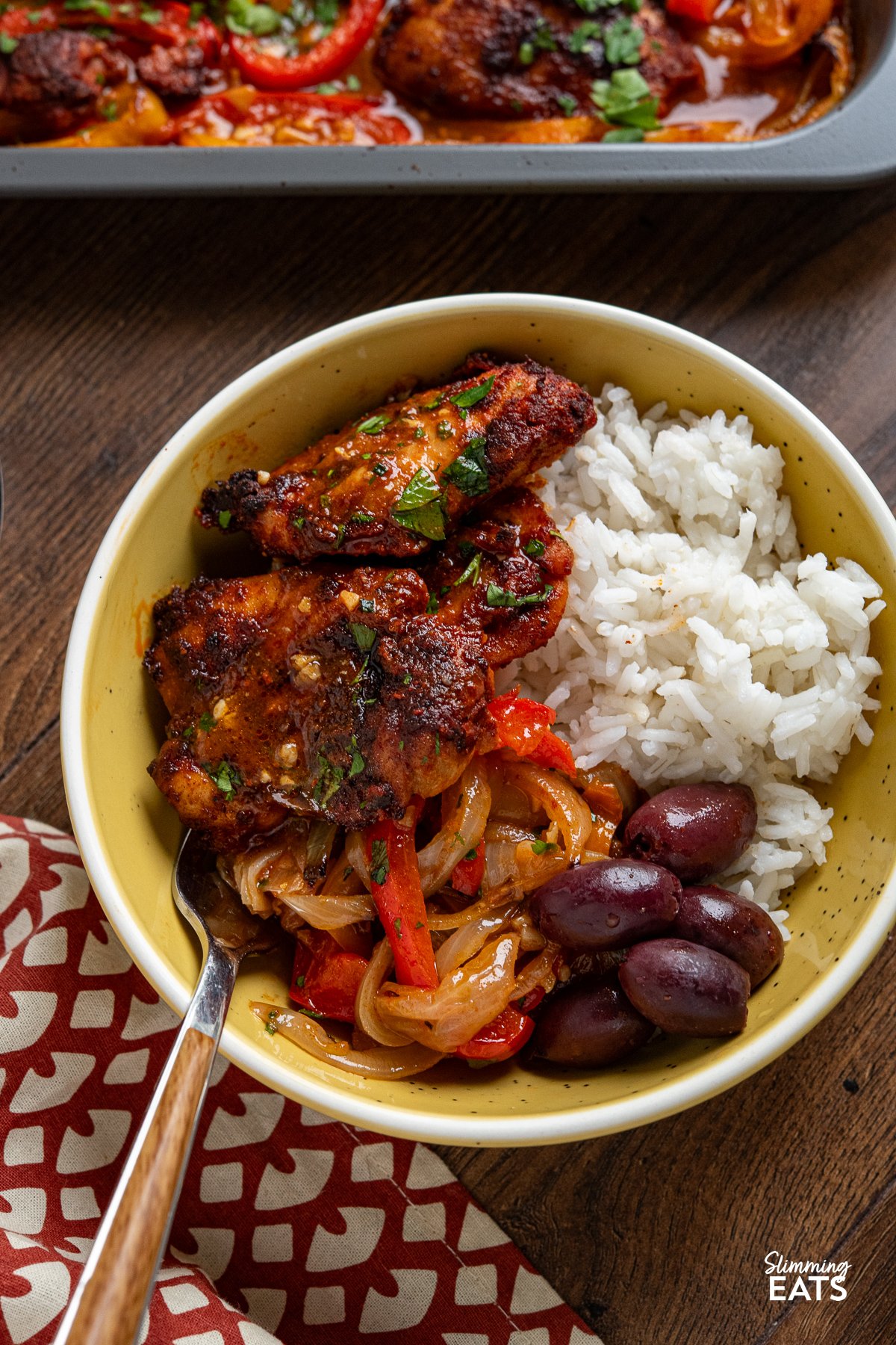 seasoned baked chicken thighs on a sheet pan with roasted juicy peppers in a yellow bowl with rice and kalamata olives