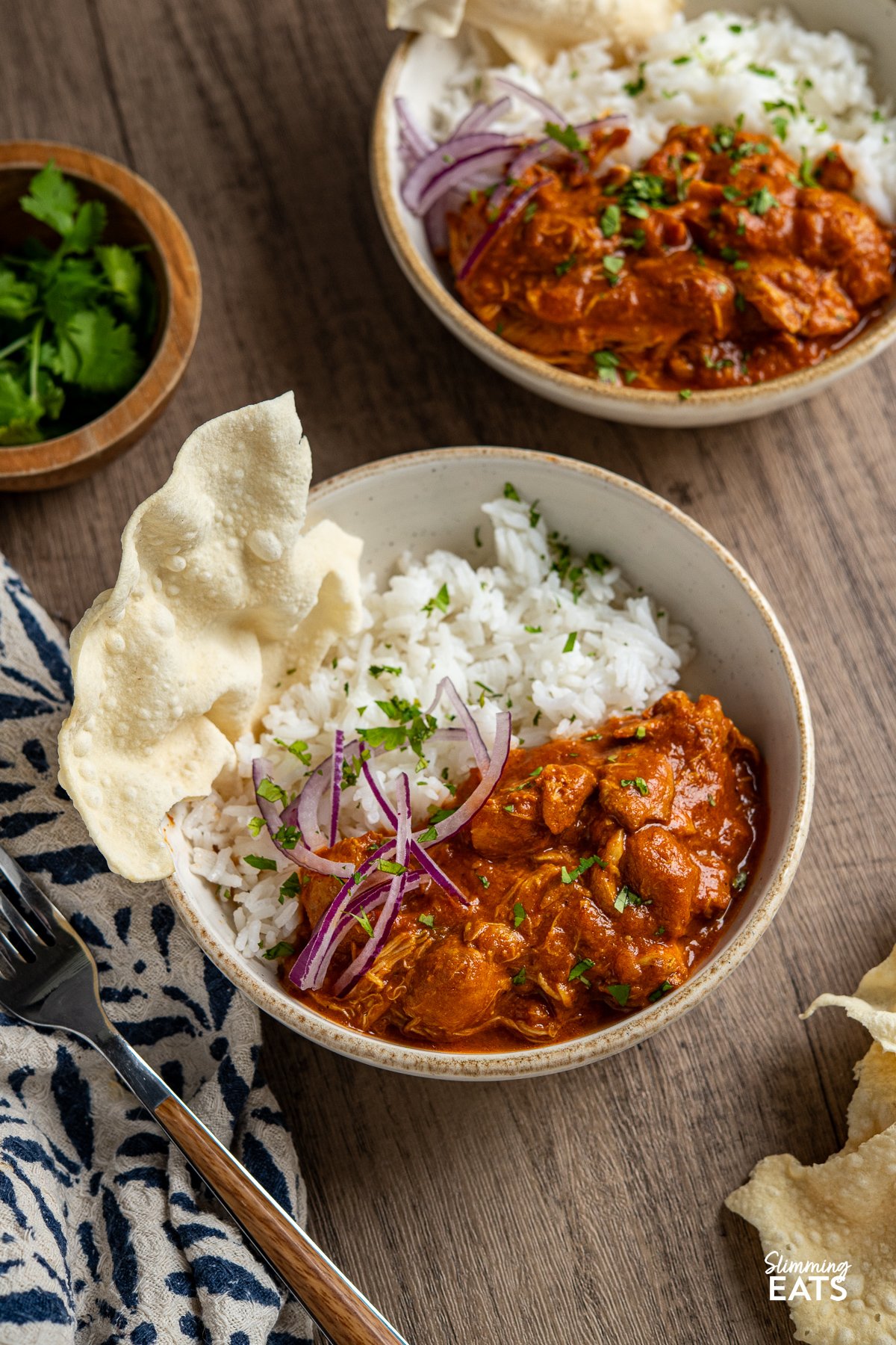 Enjoy the comforting flavours of this Easy Slow Cooker Tomato Chicken Curry. Tender chicken thighs cooked slowly in a lush tomato creamy sauce, spiced with the perfect mix of garlic, ginger, chilli and curry powder. Two white bowls with servings of slow cooker tomato chicken curry, accompanied by rice, red onion, chopped coriander, and a broken poppadum to the left.