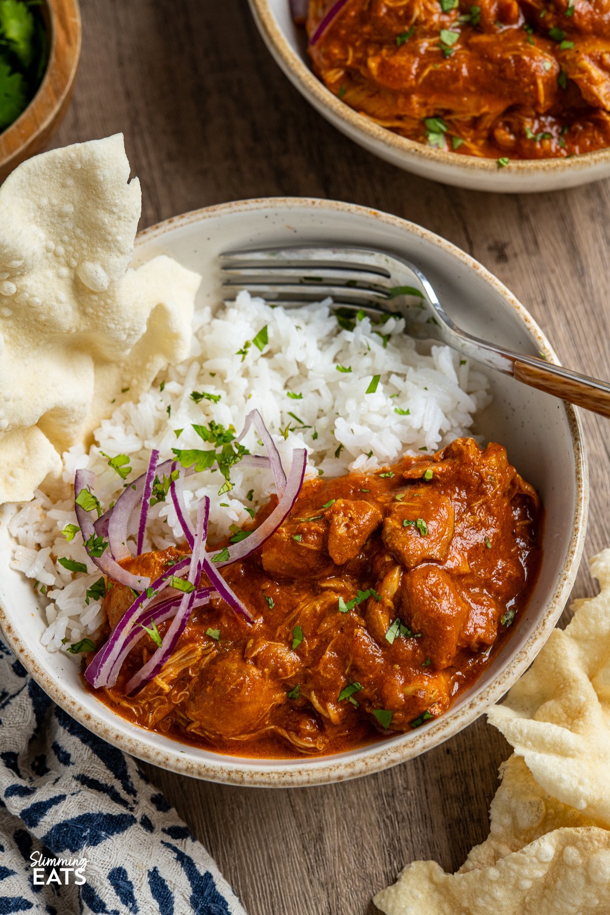 Enjoy the comforting flavours of this Easy Slow Cooker Tomato Chicken Curry. Tender chicken thighs cooked slowly in a lush tomato creamy sauce, spiced with the perfect mix of garlic, ginger, chilli and curry powder. close up of a serving of slow cooker tomato chicken curry, accompanied by rice, red onion, chopped coriander, and a broken poppadum to the left.Fork placed on side in bowl.