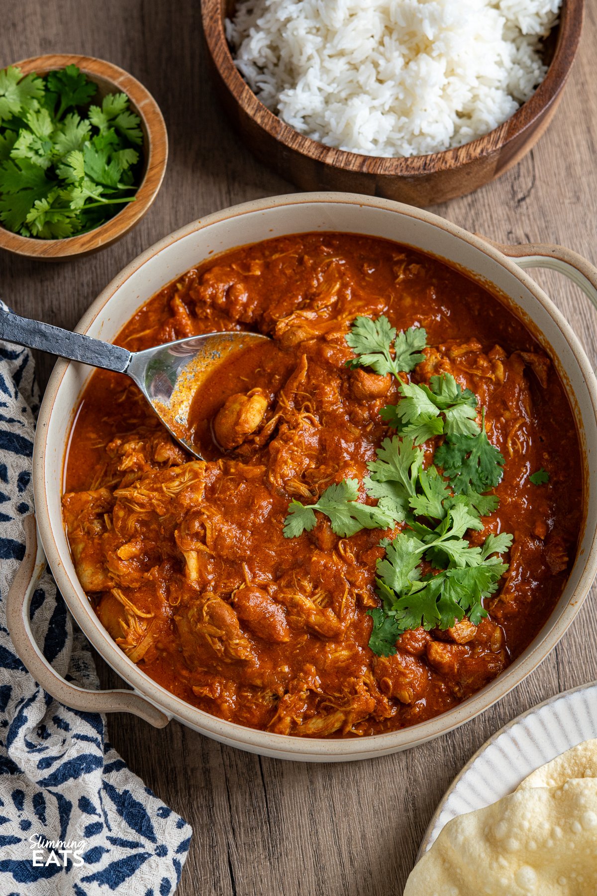 Enjoy the comforting flavours of this Easy Slow Cooker Tomato Chicken Curry. Tender chicken thighs cooked slowly in a lush tomato creamy sauce, spiced with the perfect mix of garlic, ginger, chilli and curry powder. slow cooker tomato chicken curry in a ceramic dish with spoon, wooden bowl of rice and coriander above.