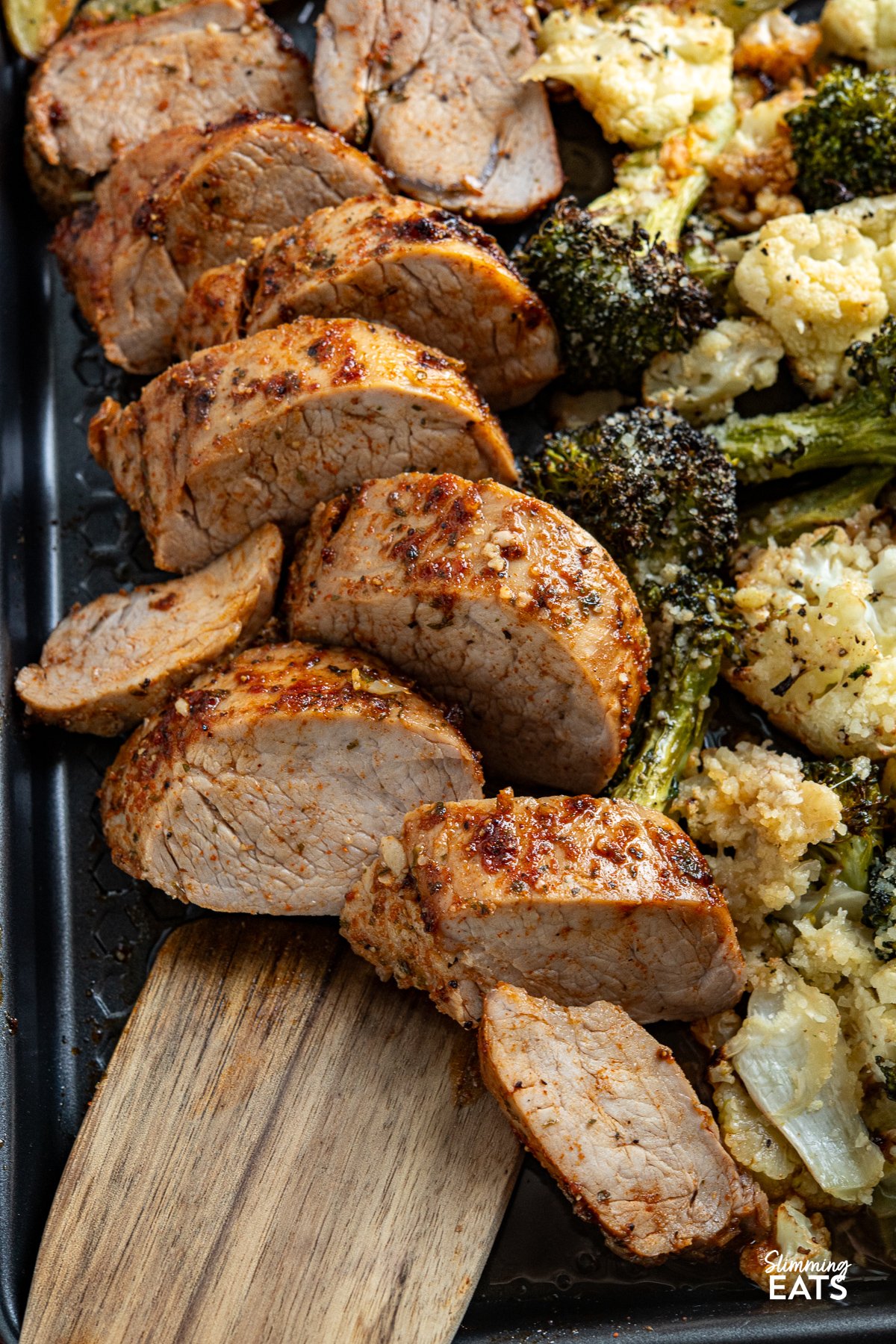 This Sheet Pan Pork Tenderloin recipe is a complete meal on one tray. Tender, juicy pork tenderloin is seasoned with a savoury, peppery spice mix and glazed with maple syrup, paired with golden, garlic-infused potato wedges and a medley of roasted broccoli and cauliflower, all topped with a cheesy sprinkle of Parmesan. Each bite offers a delightful mix of vibrant flavours and satisfying textures. close up of seasoned and pork tenderloin on a baking tray with garlic potato wedges and roasted broccoli and cauliflower, sprinkle with parmesan.