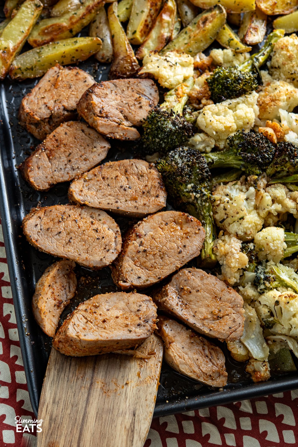 This Sheet Pan Pork Tenderloin recipe is a complete meal on one tray. Tender, juicy pork tenderloin is seasoned with a savoury, peppery spice mix and glazed with maple syrup, paired with golden, garlic-infused potato wedges and a medley of roasted broccoli and cauliflower, all topped with a cheesy sprinkle of Parmesan. Each bite offers a delightful mix of vibrant flavours and satisfying textures. close up of seasoned and pork tenderloin on a baking tray with garlic potato wedges and roasted broccoli and cauliflower, sprinkle with parmesan.